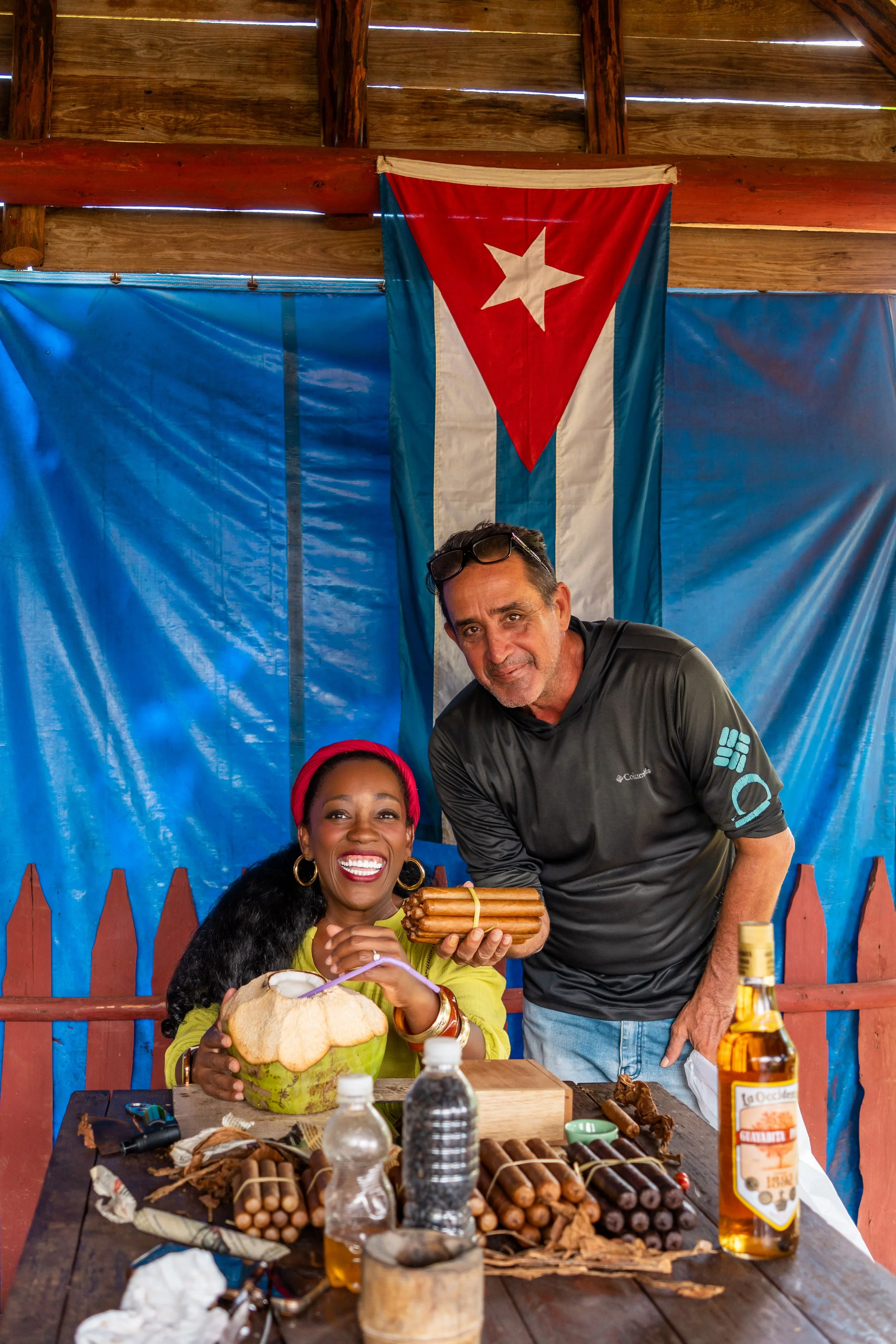 A woman with red hair and a man are sitting at a table with cigars, rum, and a coconut; the woman is holding a coconut and a bunch of cigars, smiling, while the man is leaning over and holding a bunch of cigars. A Cuban flag hangs in the background i