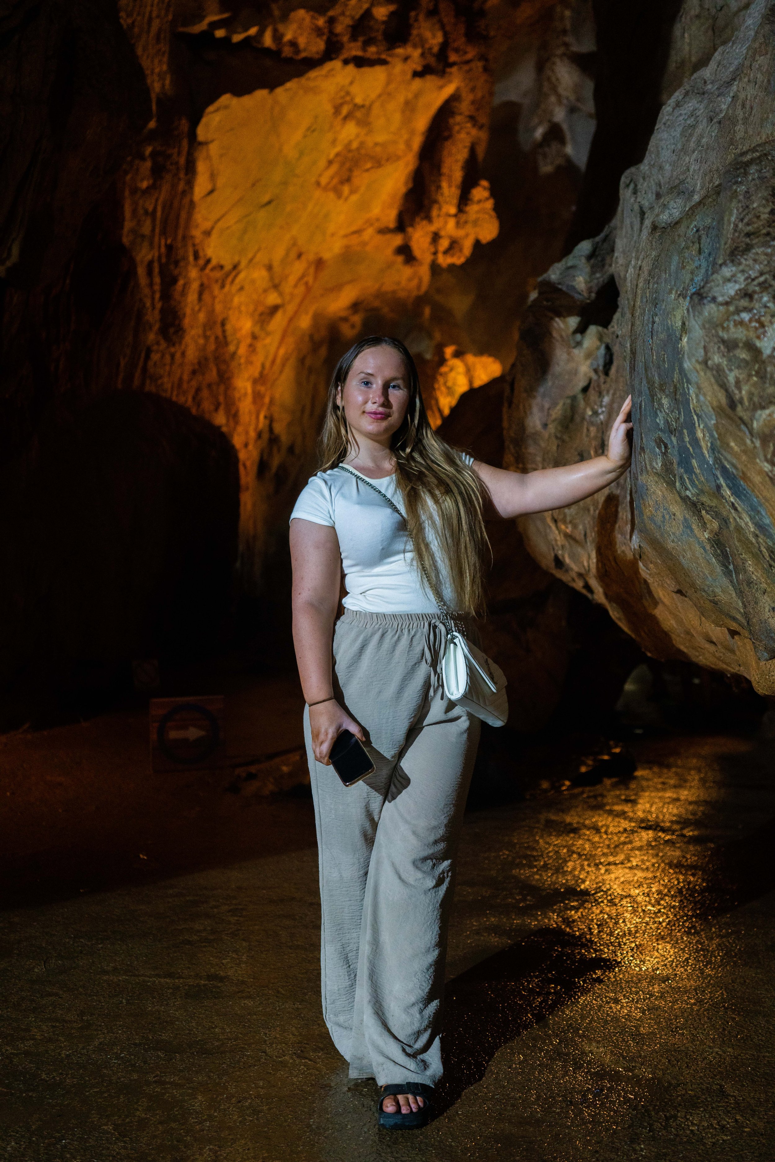 A young woman with long blonde hair standing inside a dimly lit cave, touching a rock wall with her right hand and holding a smartphone in her left hand.