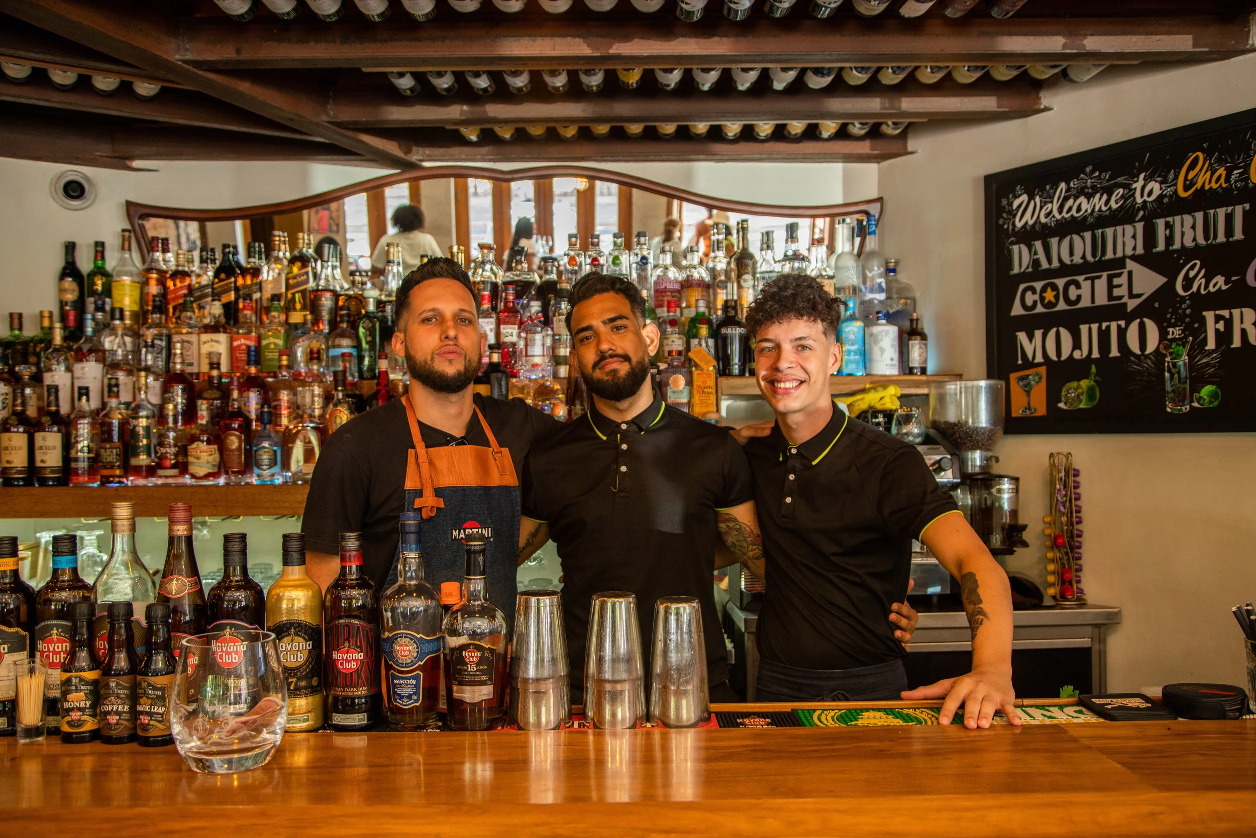 Three male bartenders behind a bar counter, smiling at the camera, with a background of numerous bottles of alcohol displayed on shelves, and a blackboard with colorful text advertising cocktails.