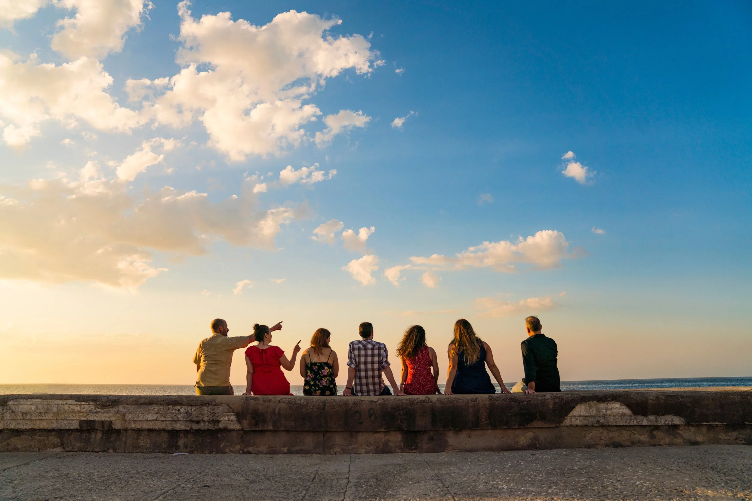 Seven people sitting on a concrete wall by the ocean at sunset, looking at the sky with clouds.