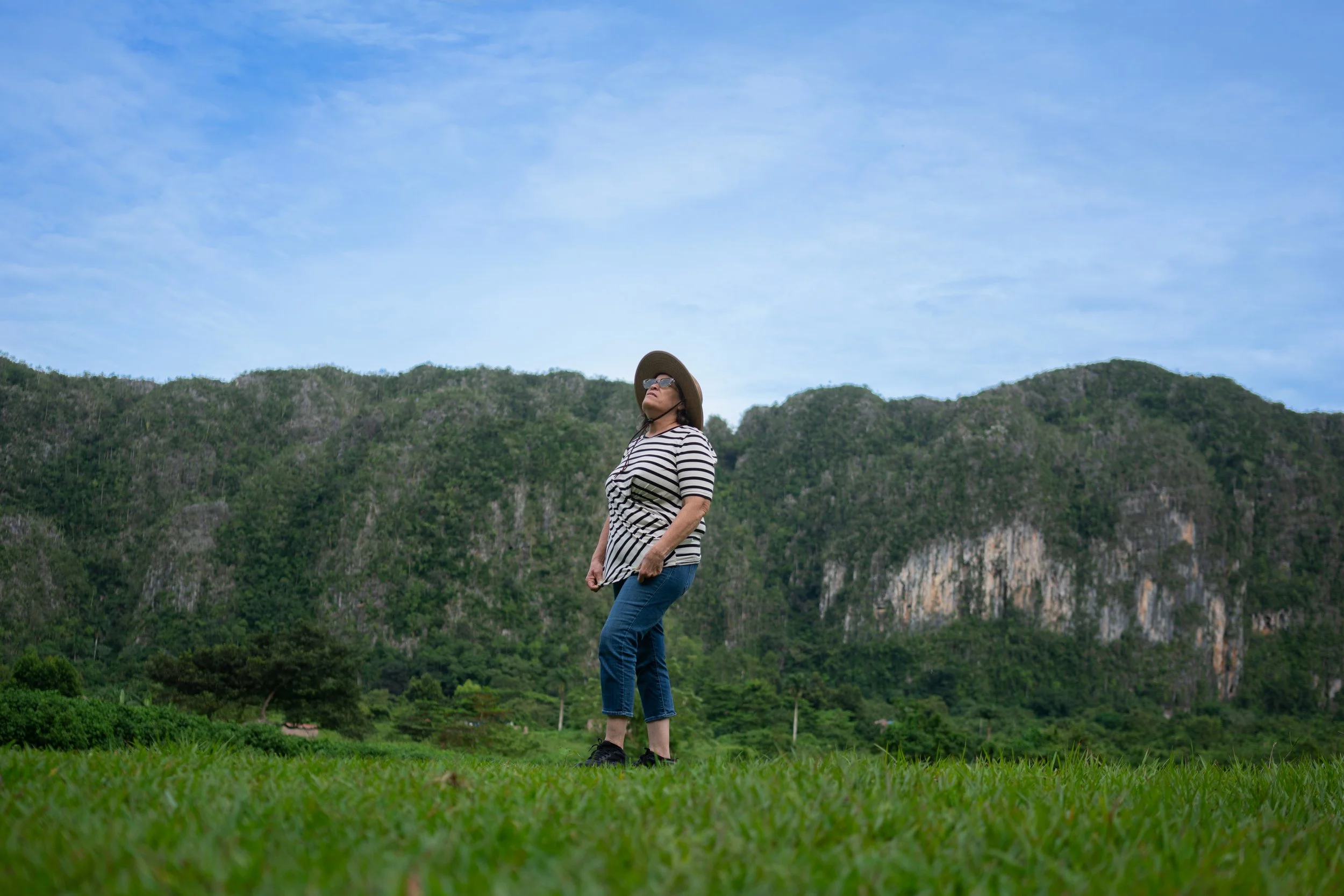A woman in a striped shirt, jeans, and a wide-brimmed hat standing on grass with green mountains in the background and a partly cloudy sky.