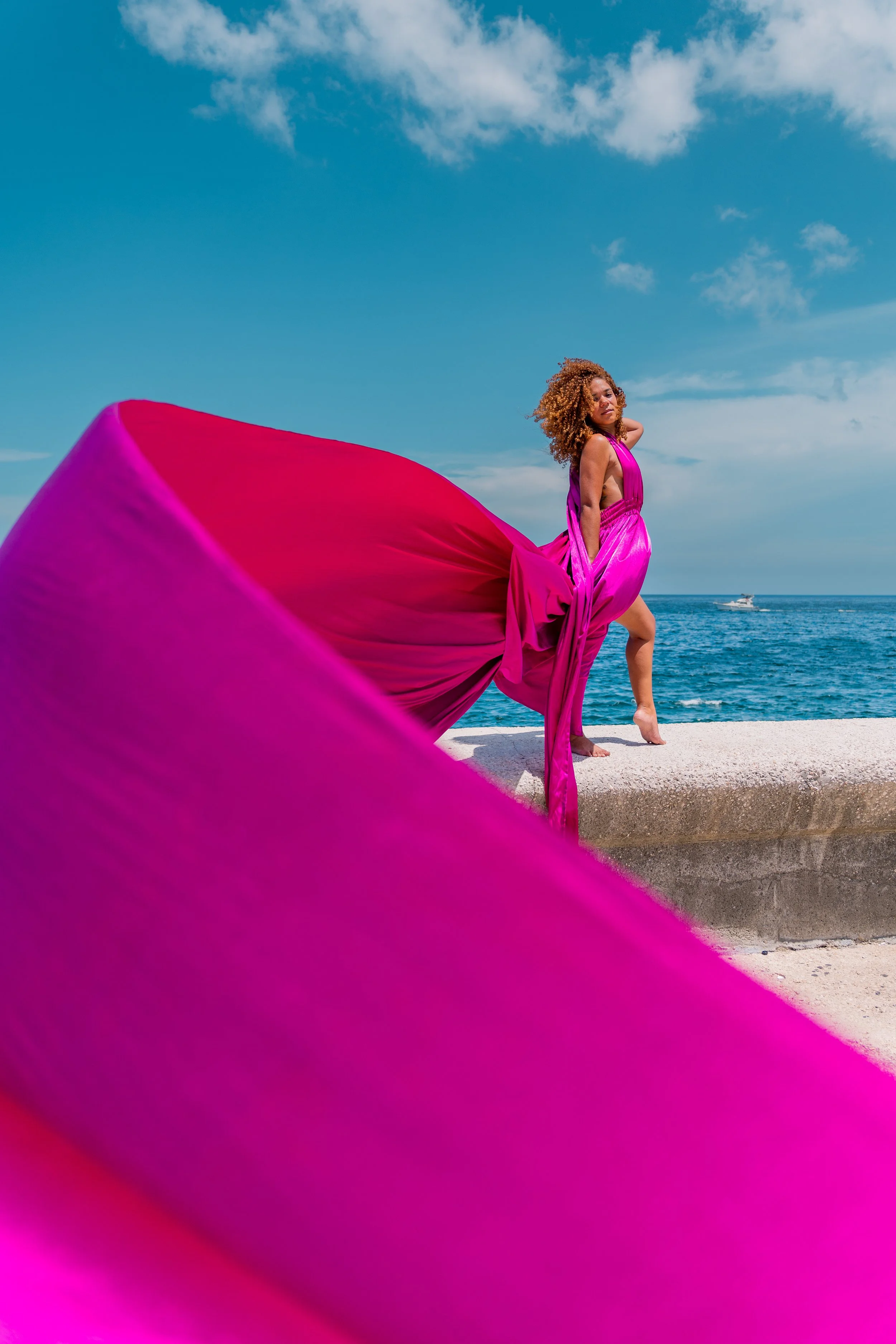 A woman in a vibrant pink dress posing near the ocean on a sunny day with blue skies and some clouds, with a boat visible in the distance.