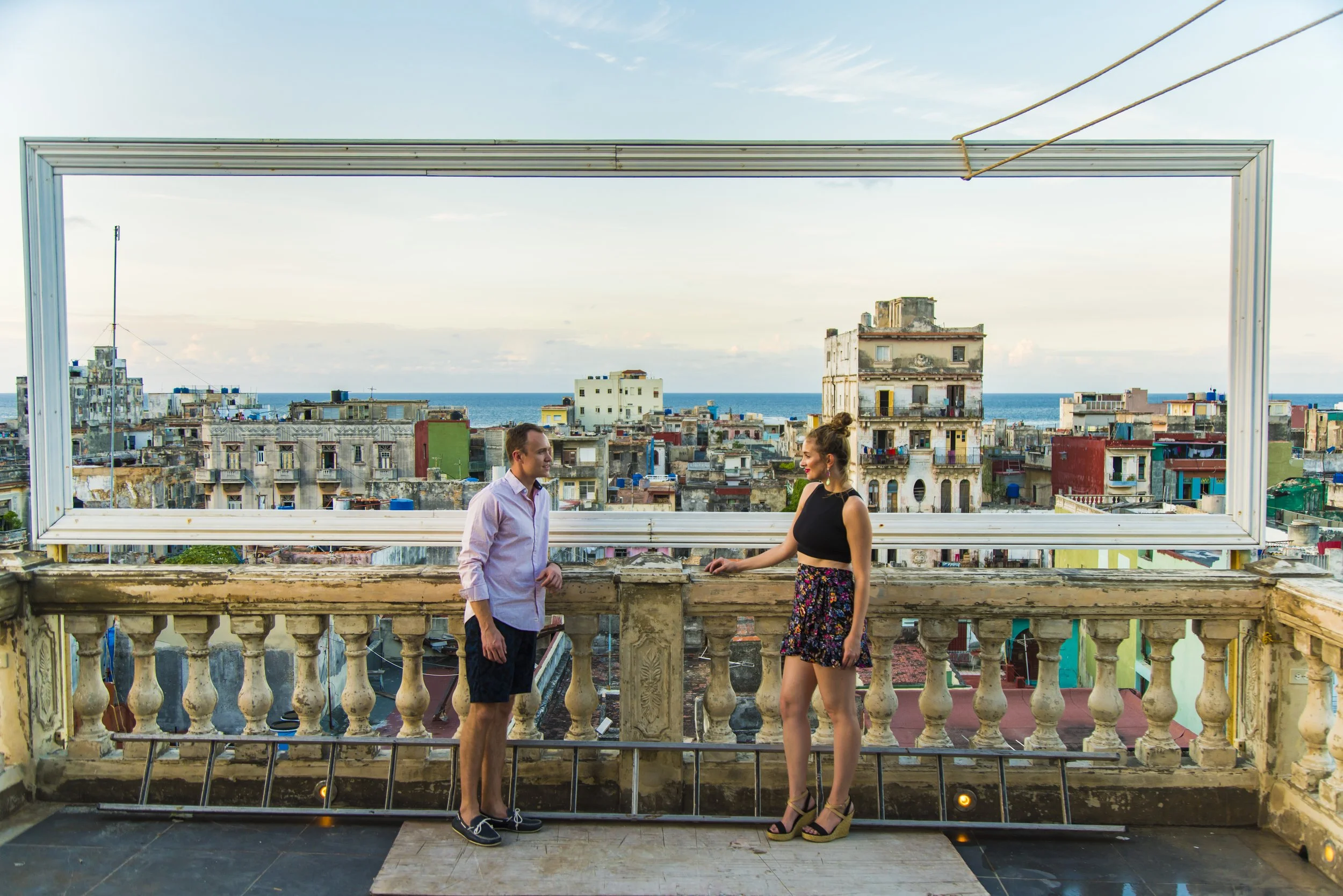 A man and woman stand on a rooftop balcony with a cityscape and ocean in the background, framed by a large rectangular window frame.