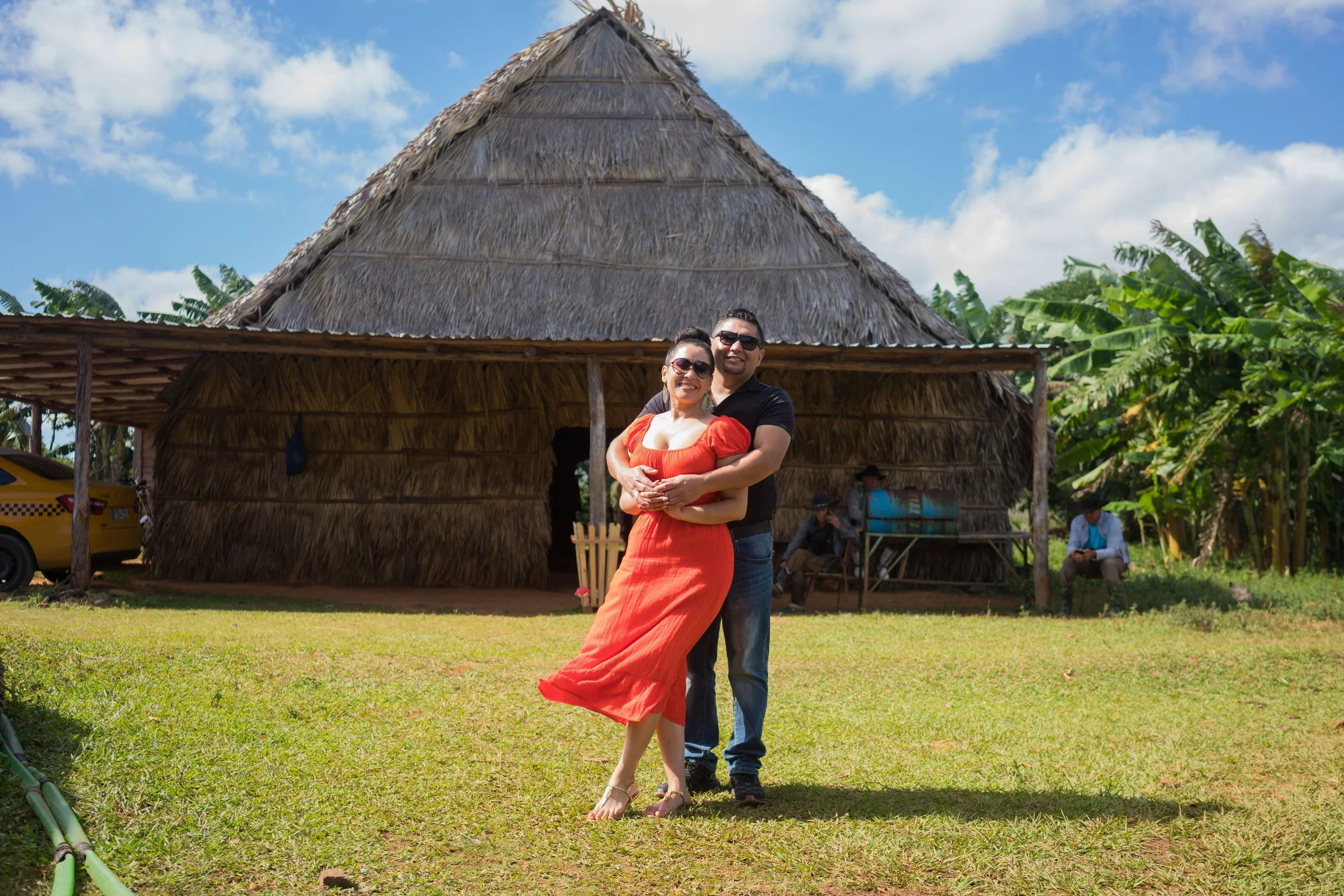 A couple hugging in front of a traditional thatched-roof hut on a sunny day, with a yellow taxi and two men sitting on a bench nearby.