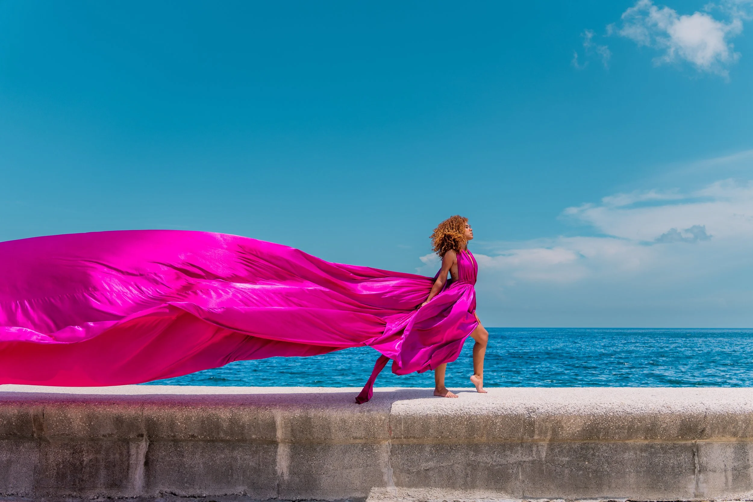 A woman in a flowing pink dress standing on a concrete ledge by the ocean with a blue sky overhead.