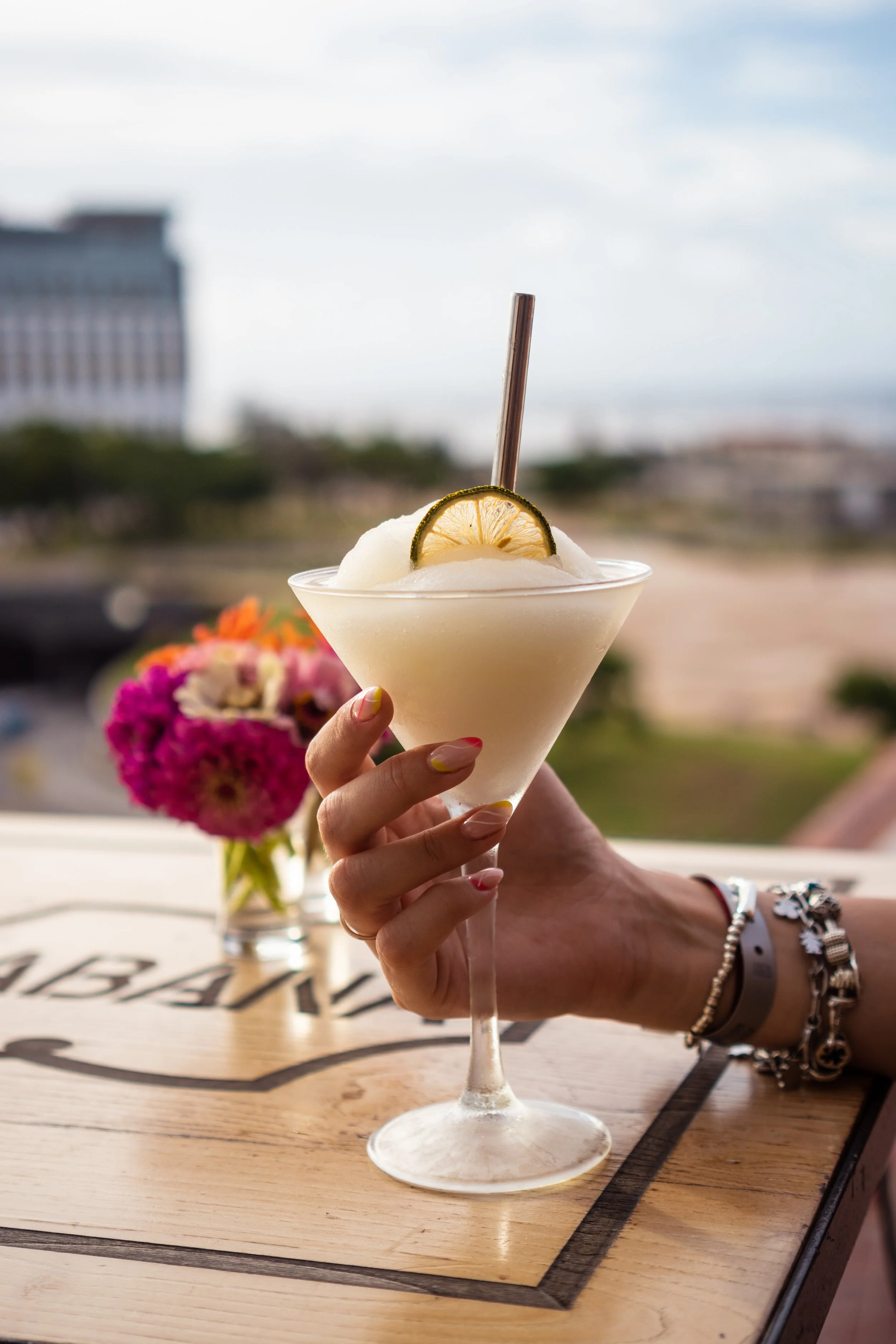 Person holding a cocktail glass with a lemon slice garnish, ice, and a straw, with a vase of colorful flowers in the background.