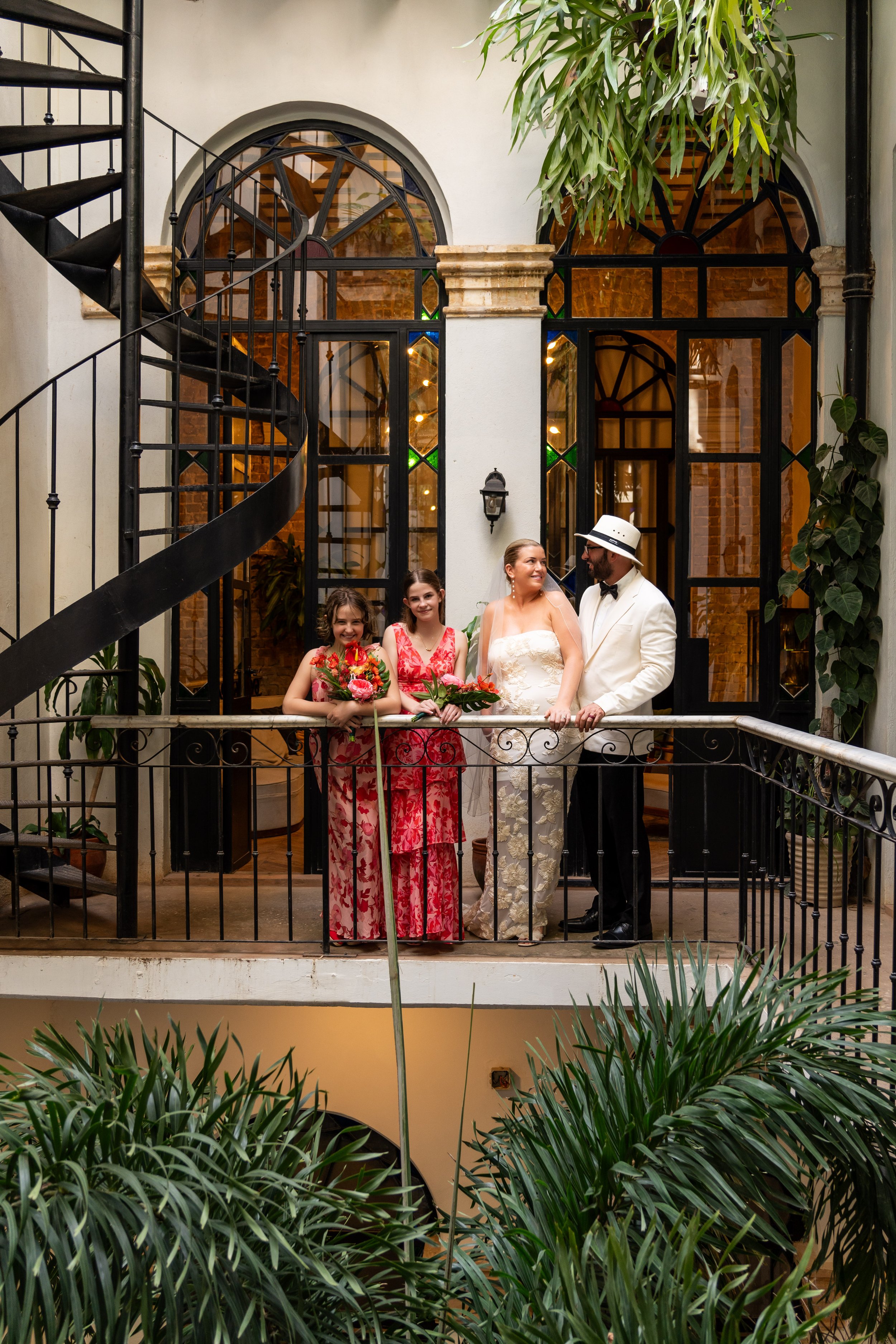 A group of four people, including a bride, groom, and two young girls, standing on a balcony in an indoor space with large windows and greenery.