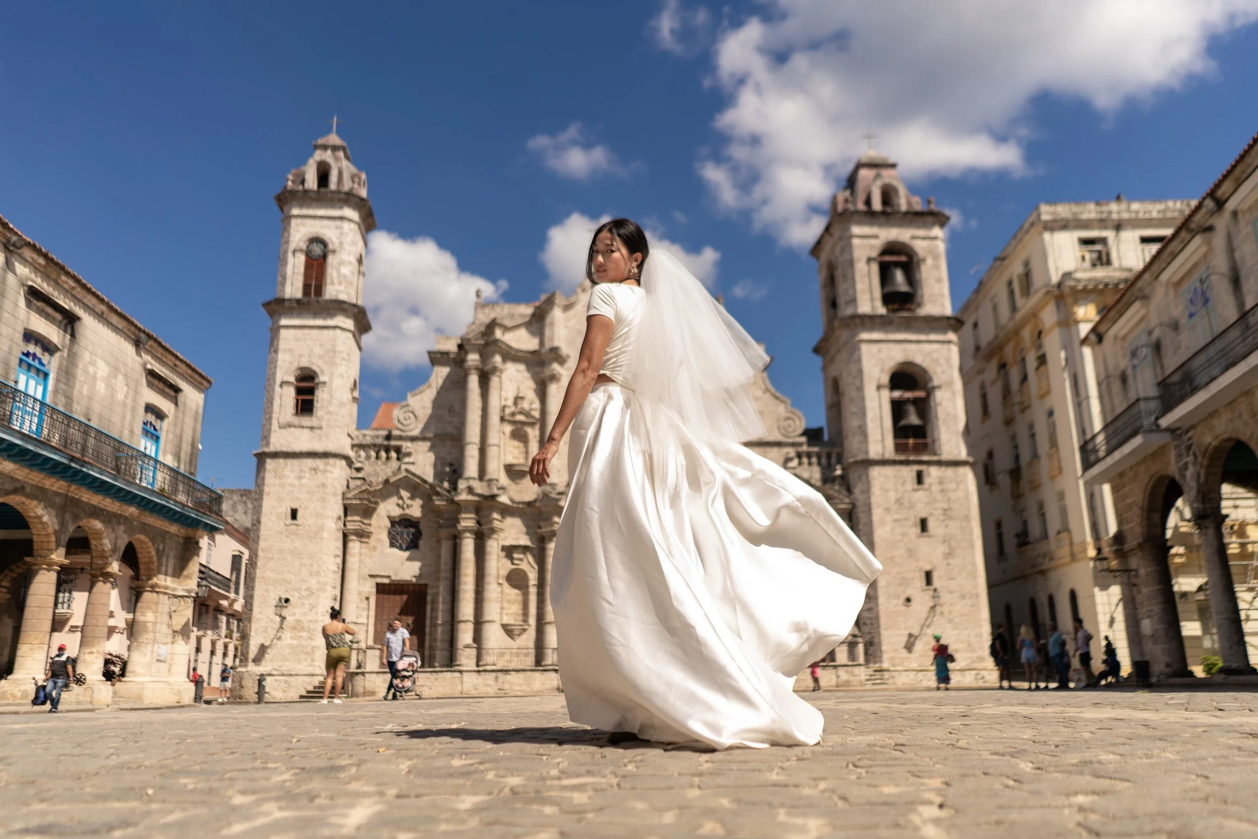A bride in a white wedding dress and veil standing outdoors in front of an old stone church, with a blue sky and clouds overhead.