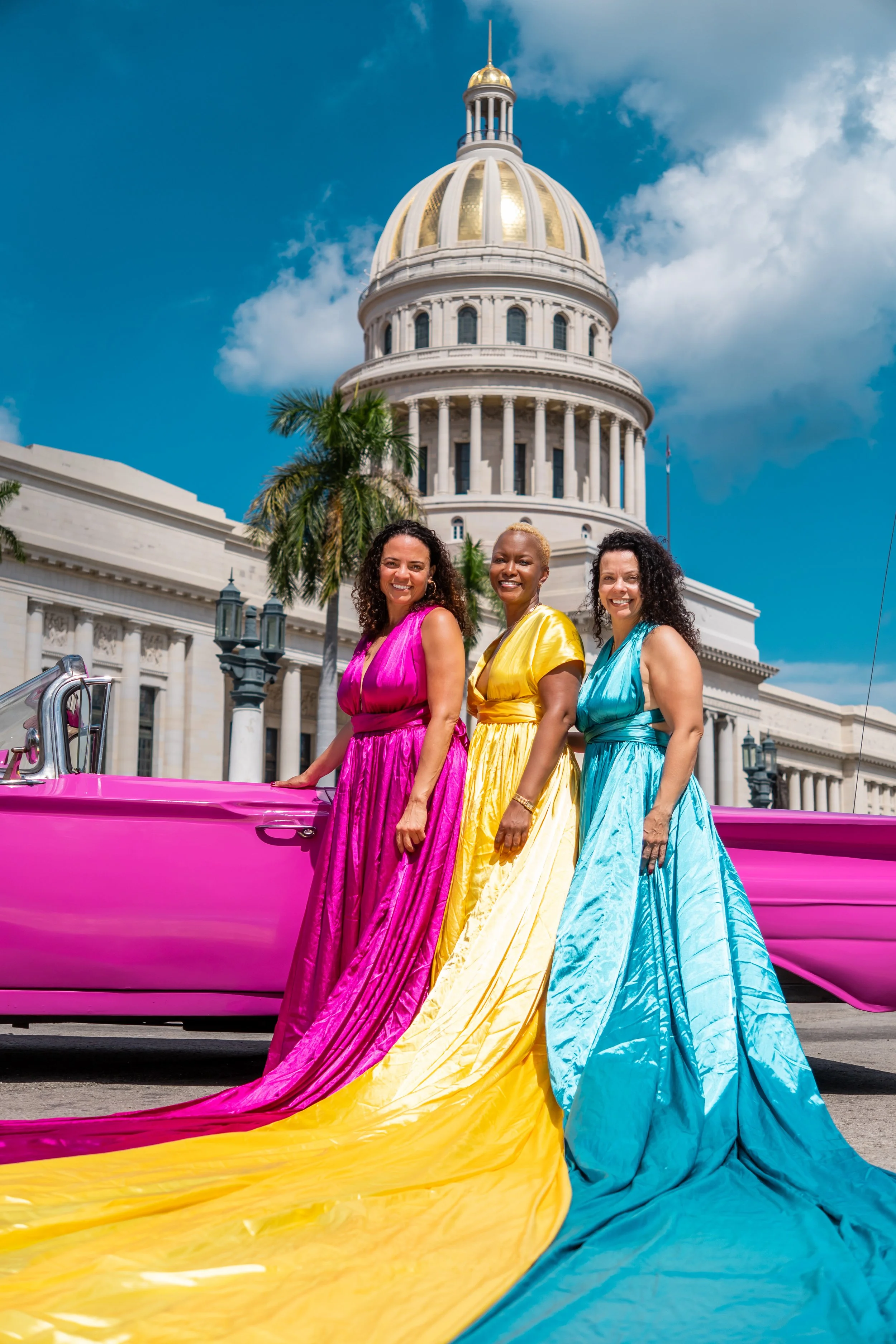 Women in colorful dresses standing in front of a pink vintage car and the Capitol building in Havana.