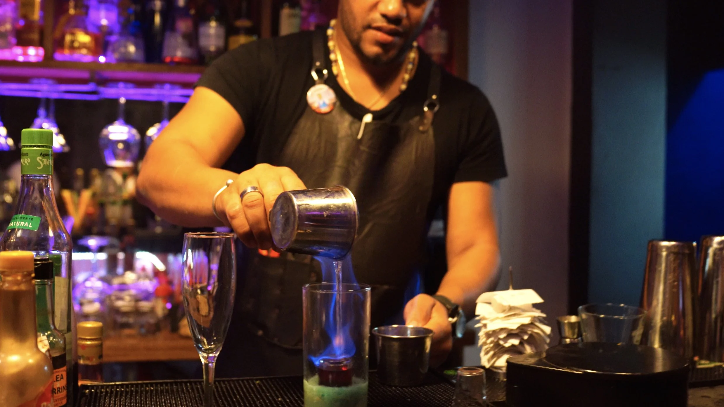 Barmans pouring a drink into a tall glass with a small flame underneath, surrounded by various bottles and bar tools.