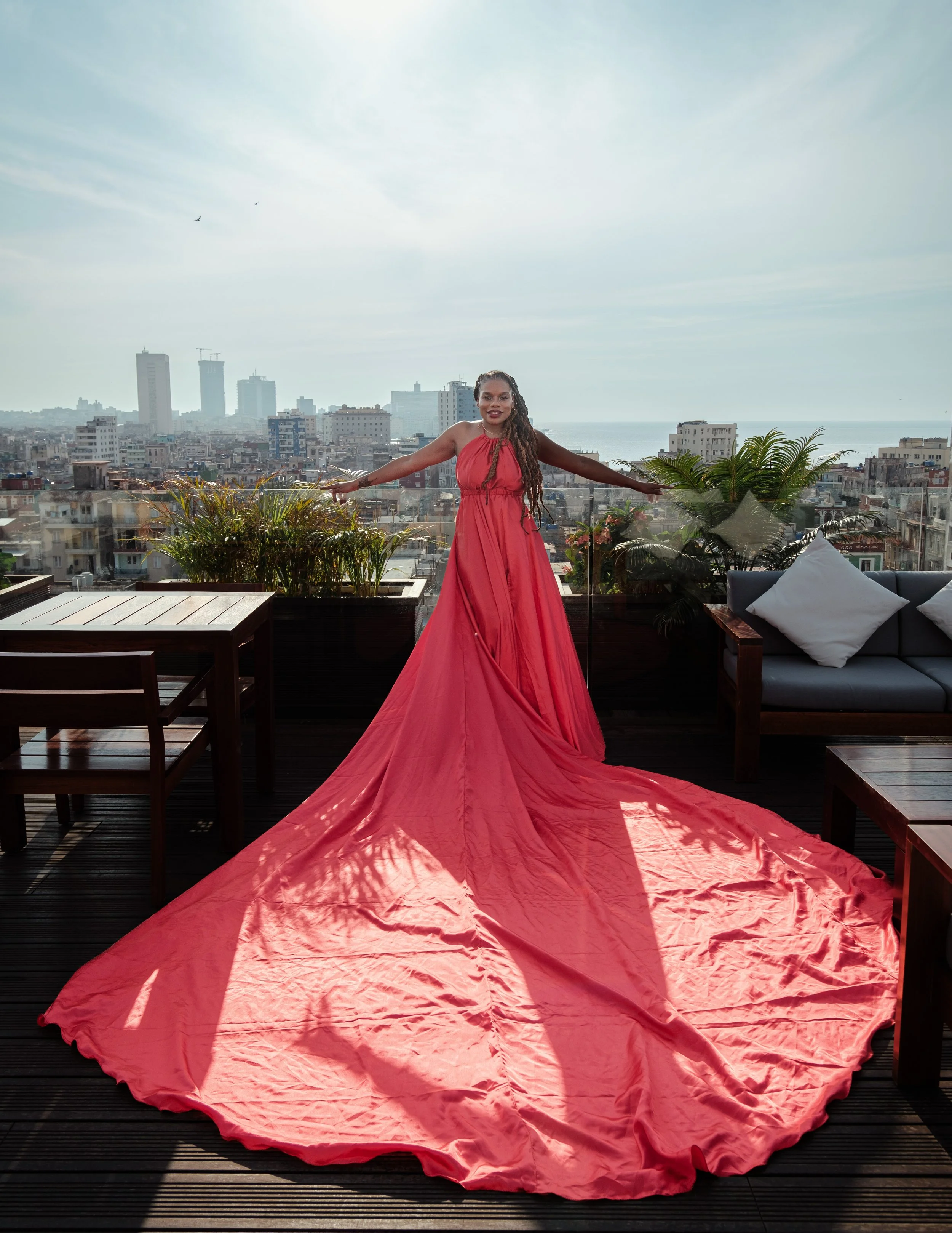 Woman in a long red gown standing on a rooftop terrace with cityscape in the background, arms outstretched, and the dress's train flowing on the wooden floor.