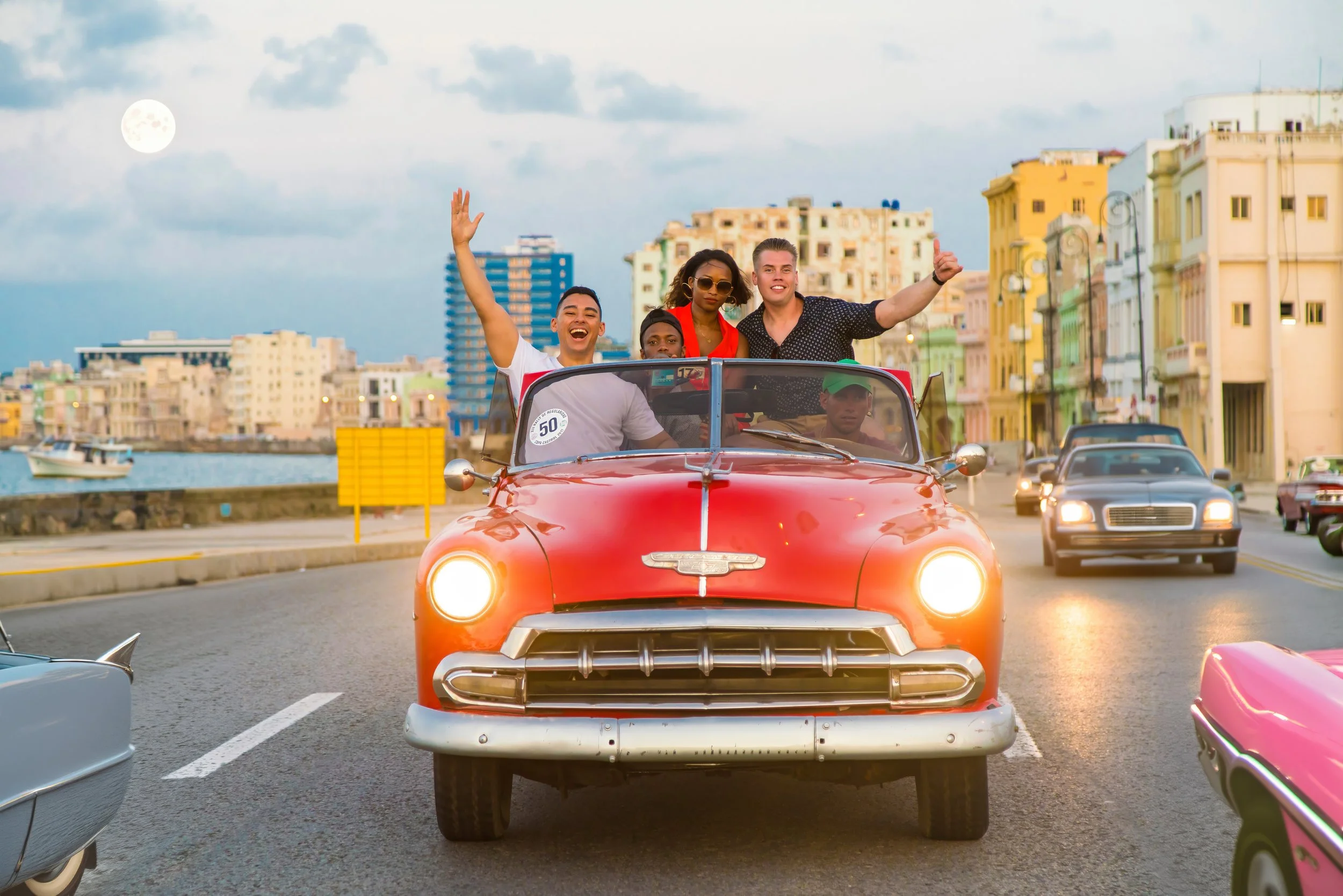 Five ethnically diverse young people riding in a red vintage convertible car with the top down on a city street during early evening, smiling and enjoying the ride.