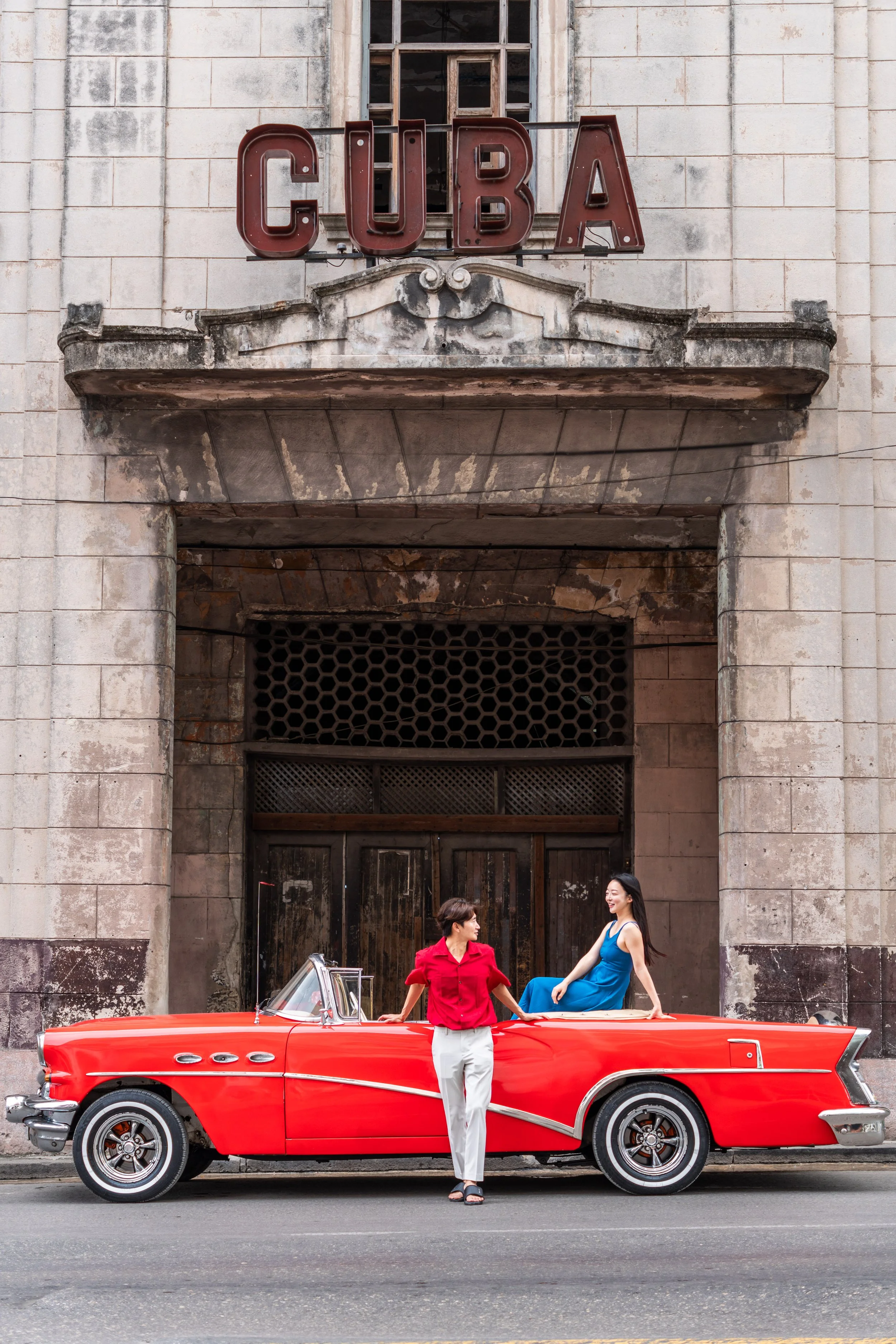 Two women and a red vintage convertible car parked in front of an old building with a sign that reads 'CUBA'.