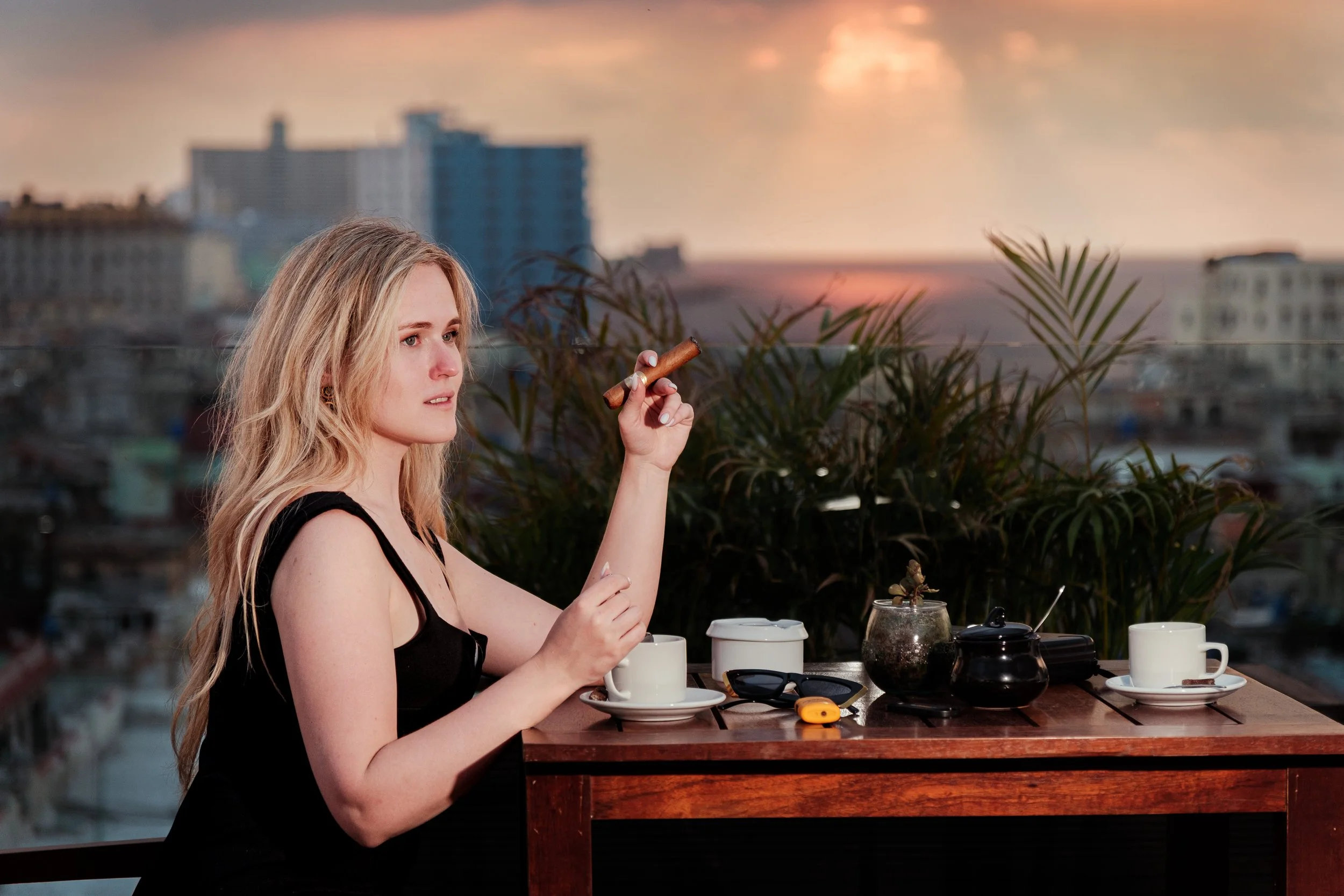 A woman with long blonde hair sitting at a wooden table on a rooftop, holding a cigar. The table has two cups, sunglasses, and small containers. In the background, there is a city skyline at sunset with a cloudy sky.