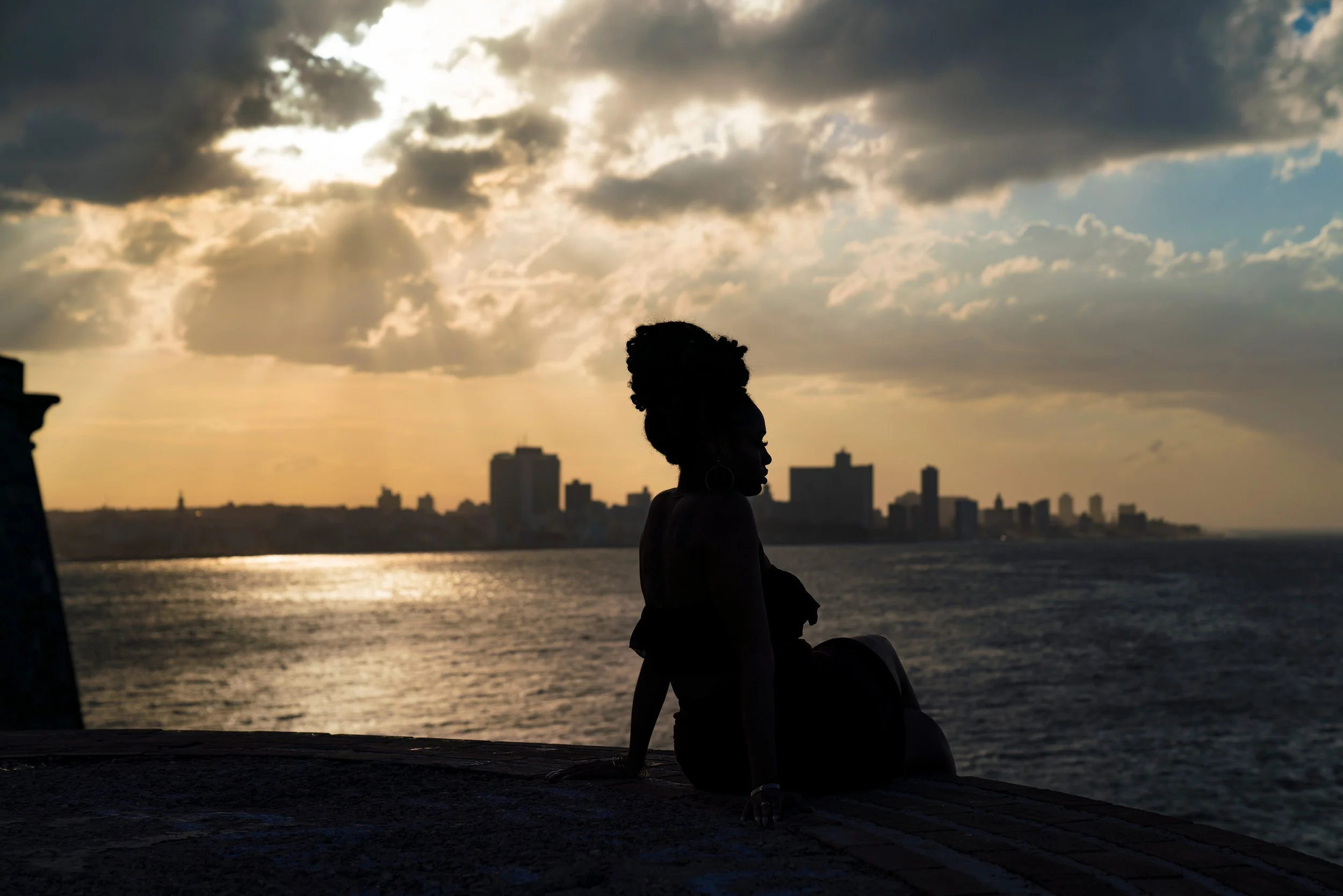 Silhouette of a woman sitting on a ledge overlooking the ocean during sunset, with a city skyline in the background.