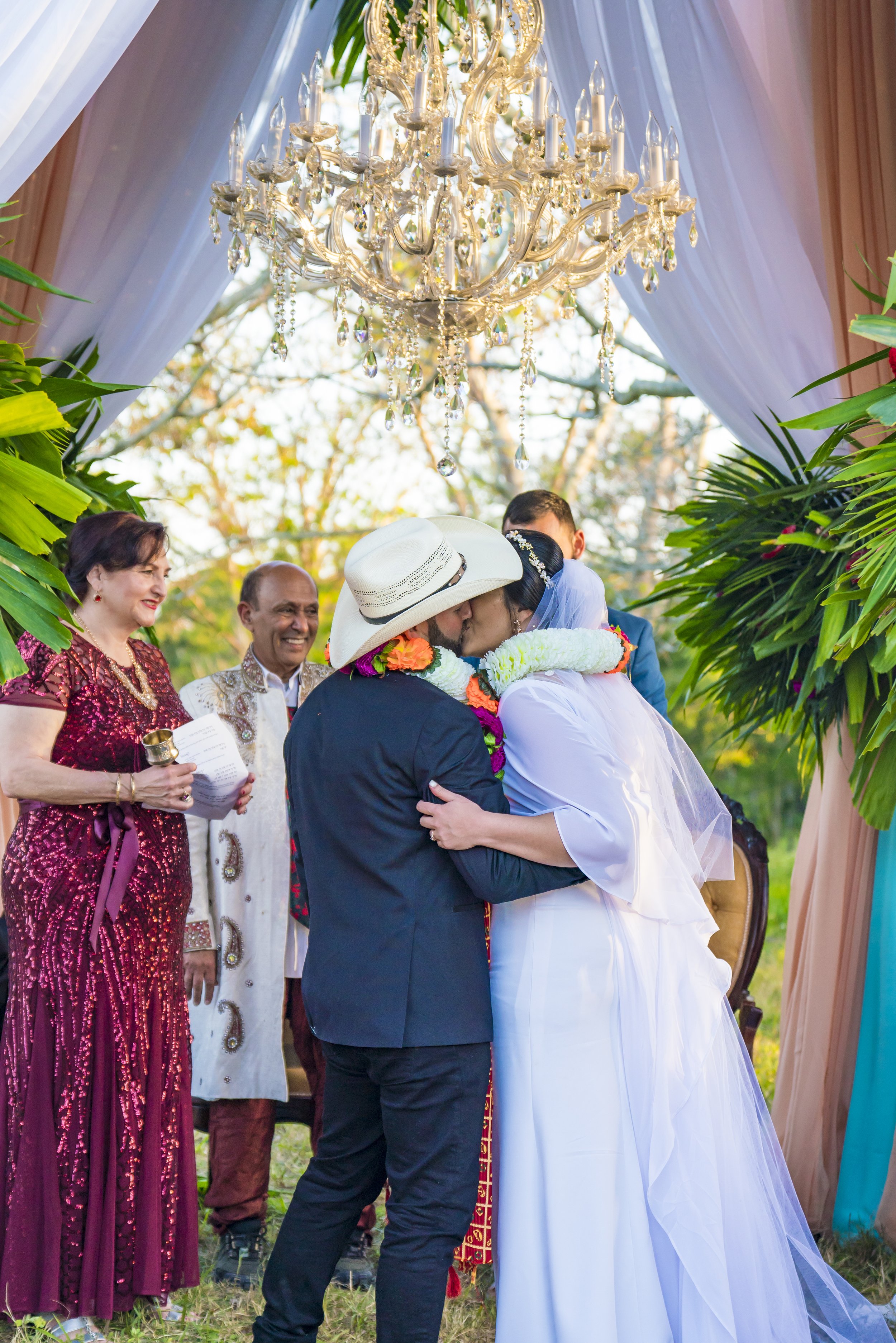 A wedding ceremony with a bride and groom kissing, surrounded by friends and family, outdoors under a chandelier with white drapes and lush green plants.