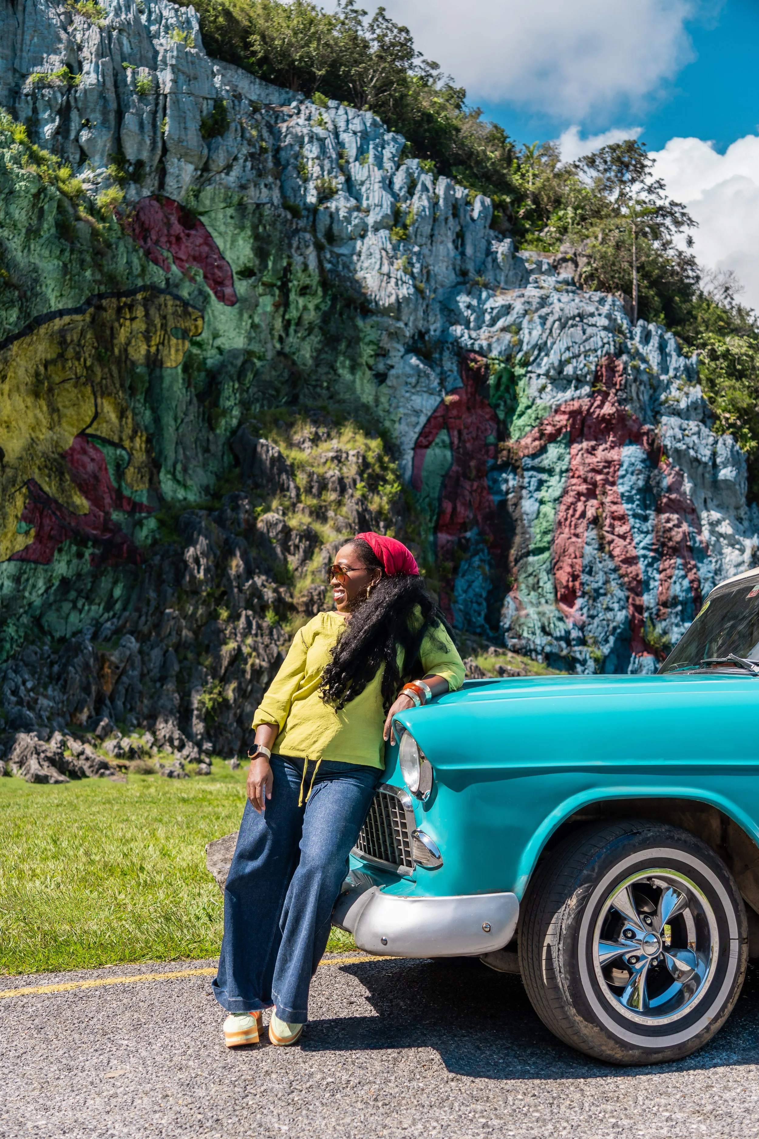 A woman with long black hair, sunglasses, yellow top, and blue jeans leaning against a turquoise vintage car on a mountain road. She is smiling and standing in front of a mountain with colorful painted figures.
