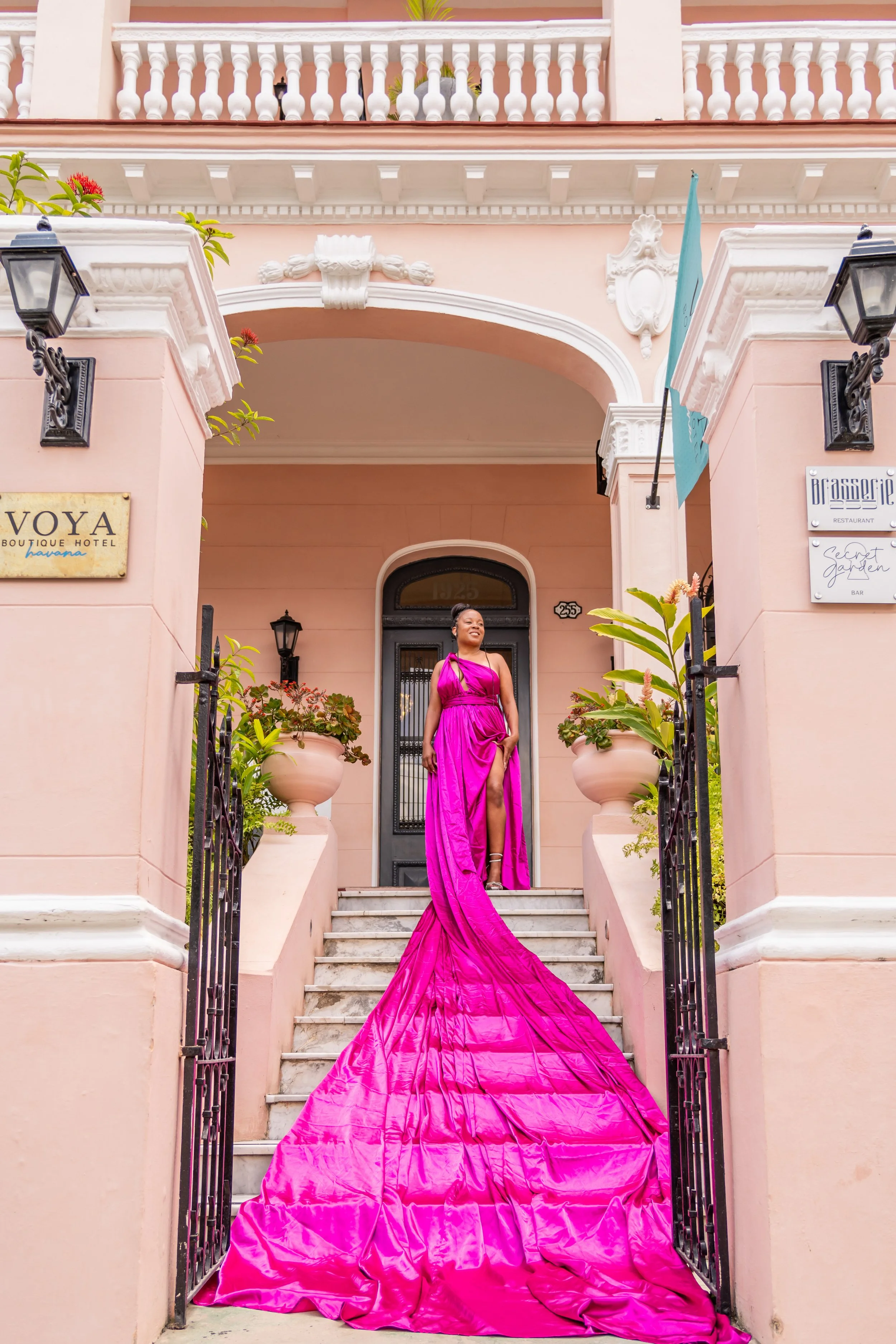 A woman in a vibrant pink gown standing on the steps of a pastel pink building, smiling.