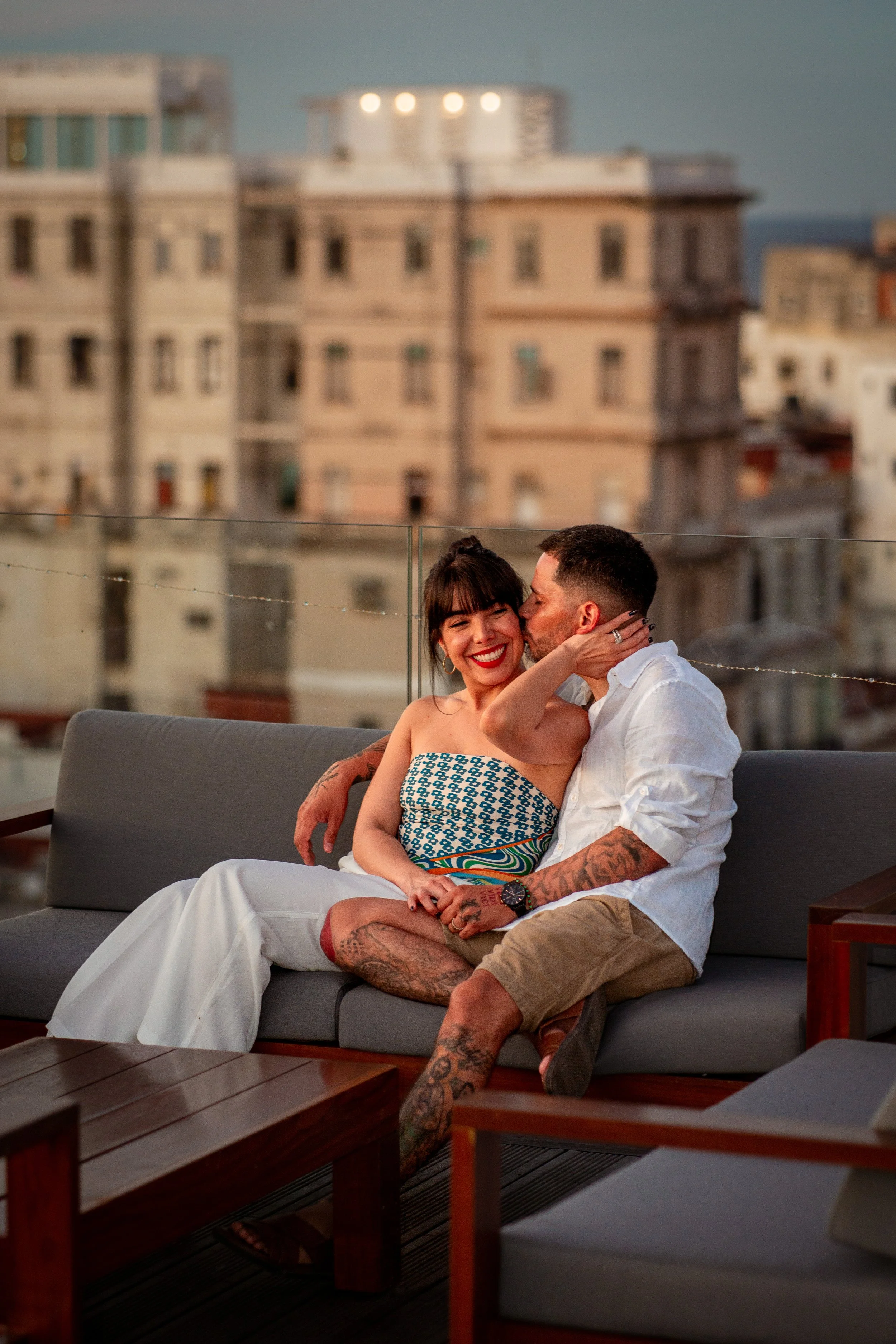 A couple sitting on an outdoor sofa at sunset, with city buildings in the background. The woman is smiling and the man is kissing her cheek.