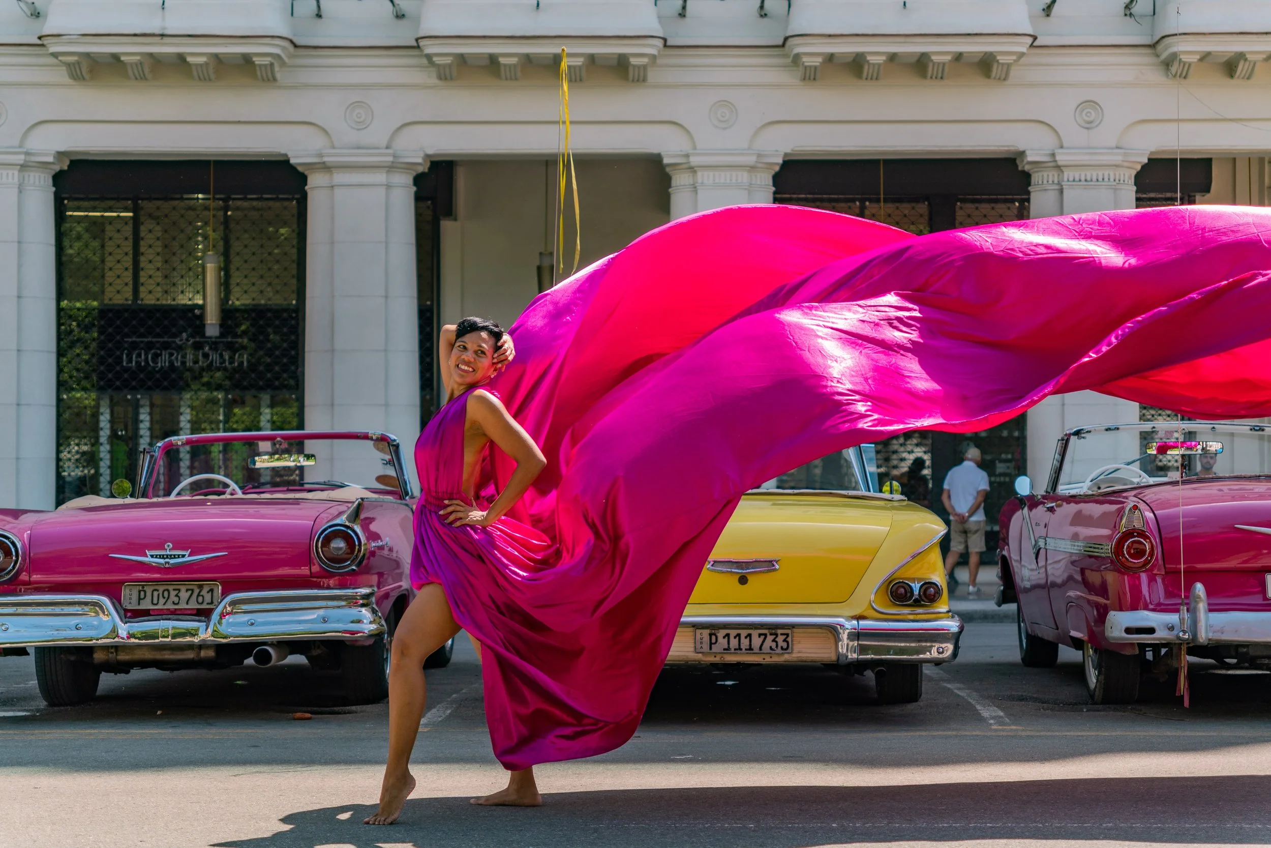 A woman in a pink dress dancing barefoot on the street in front of vintage cars with a large pink fabric flowing behind her.