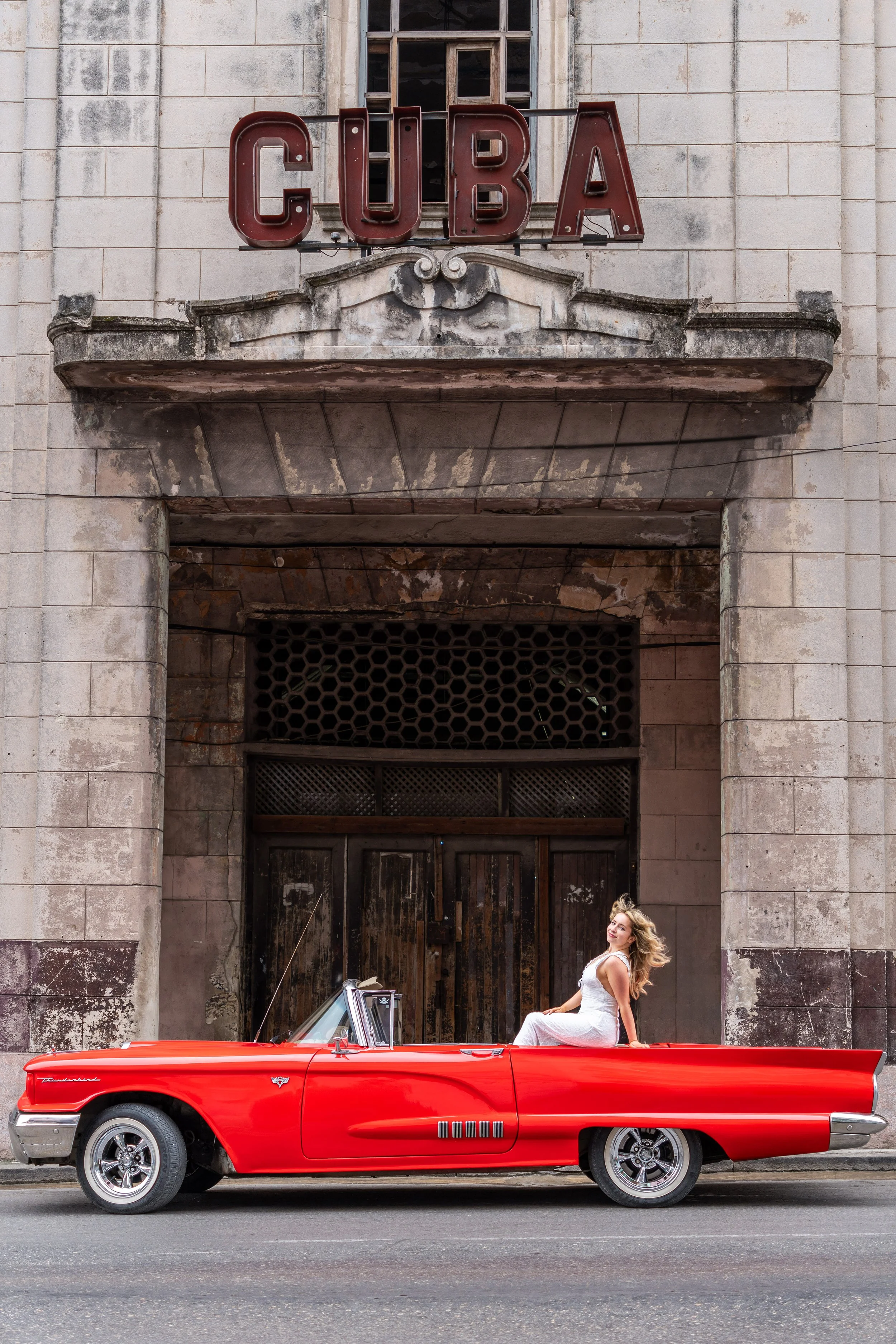 Woman sitting on a vintage red convertible parked in front of an old building with a 'CUBA' sign.