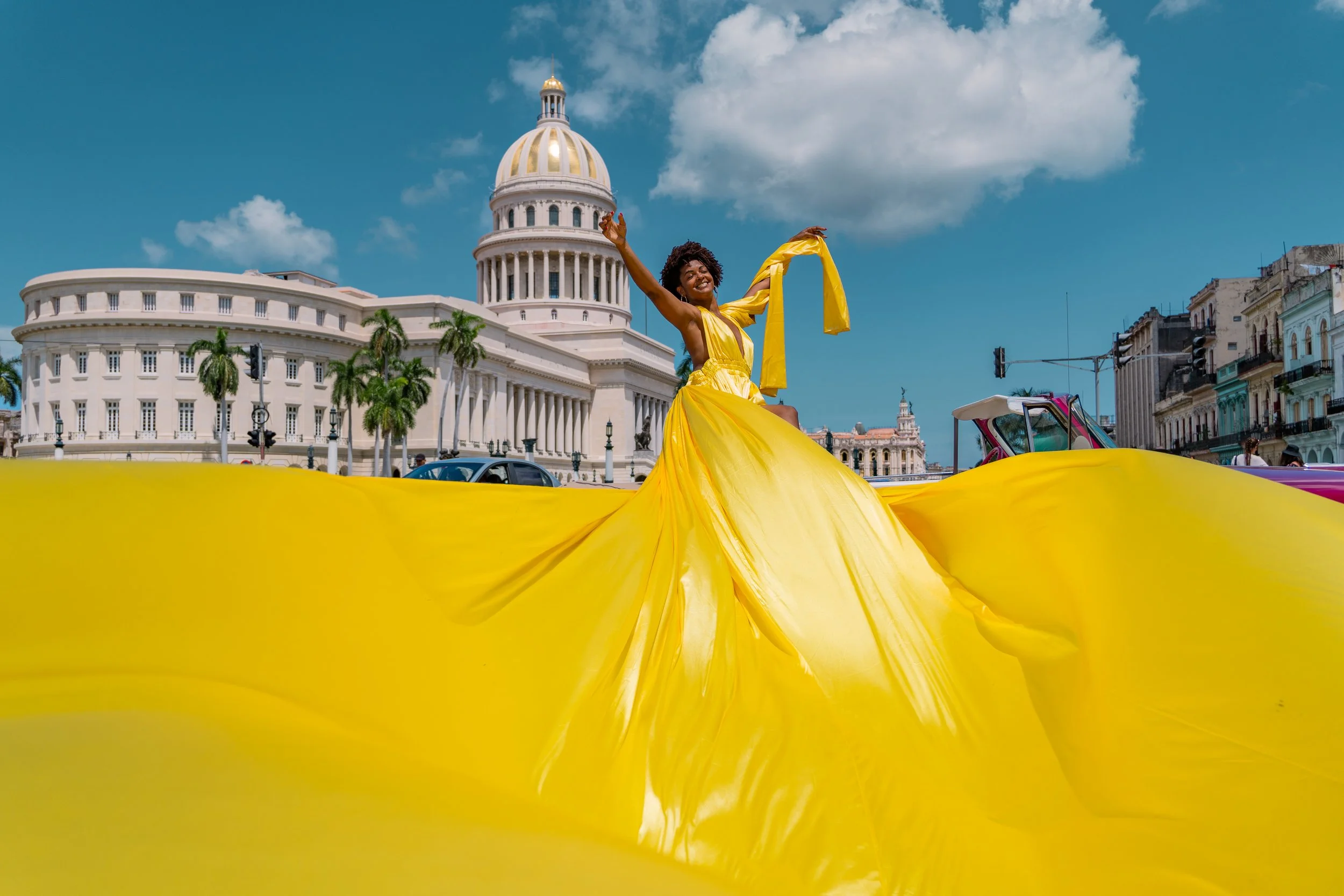 A woman in a flowing yellow dress dancing with arms raised in front of a government building with a large dome, palm trees, and a city street with cars and buildings.
