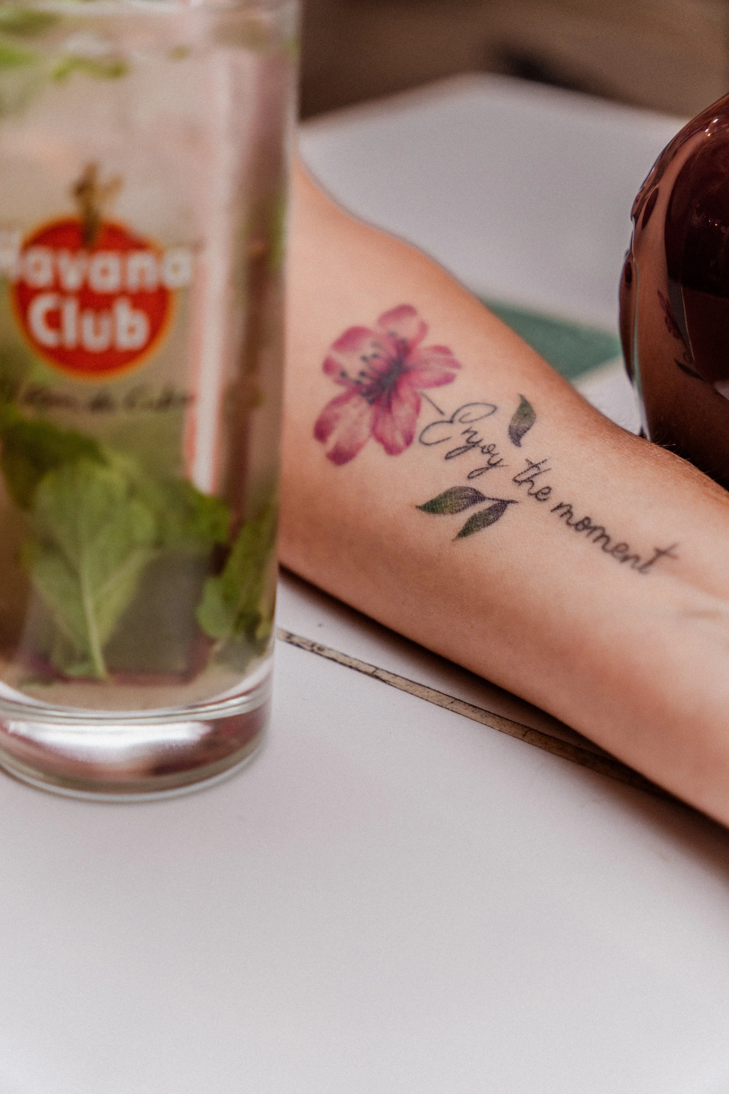 A glass of iced tea with mint leaves, a notebook with a pink flower tattoo and the words "Enjoy the moment," and a dark-colored spherical object on a white surface.