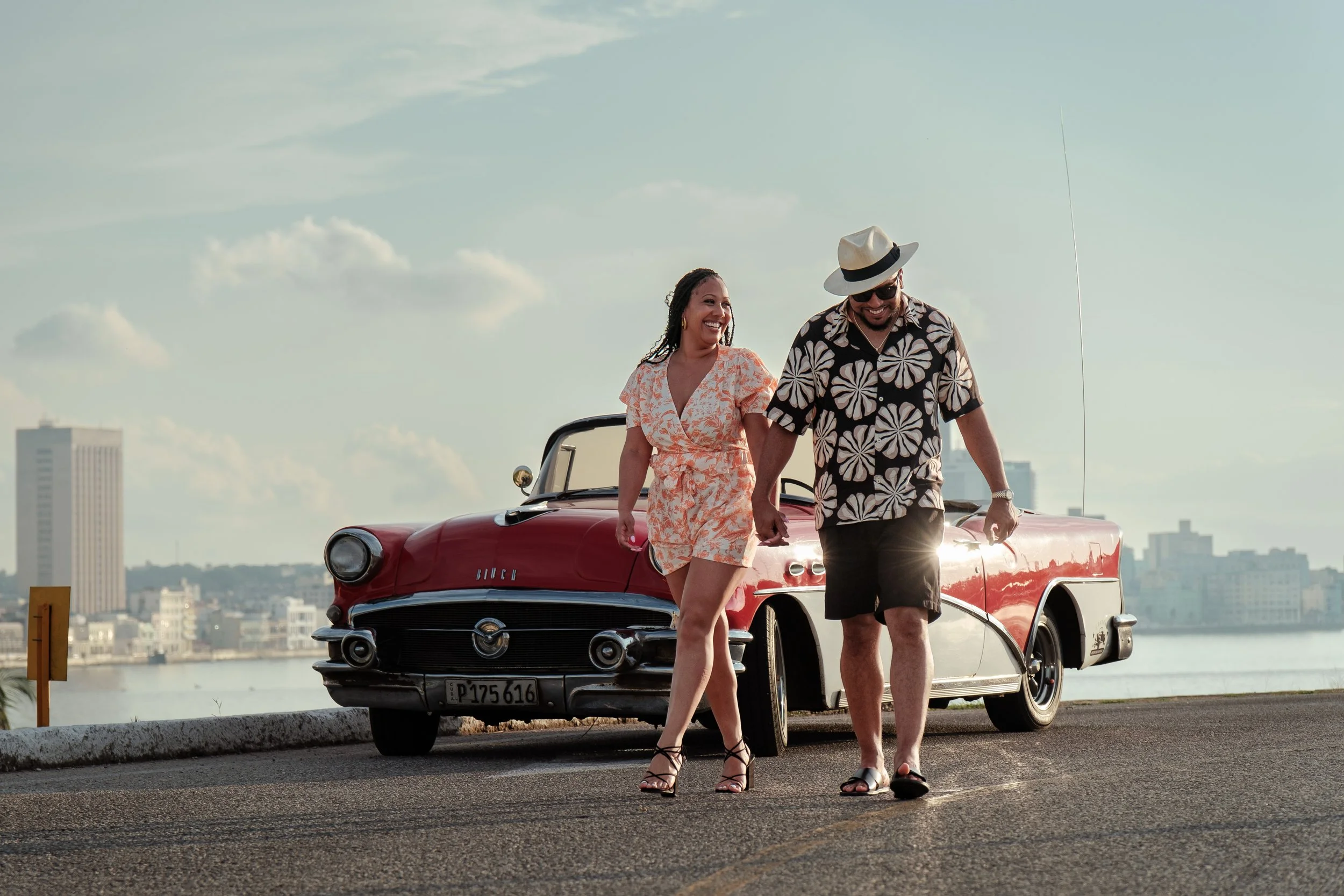 A smiling woman and man walking hand in hand by the water near a vintage red Cadillac during sunset.