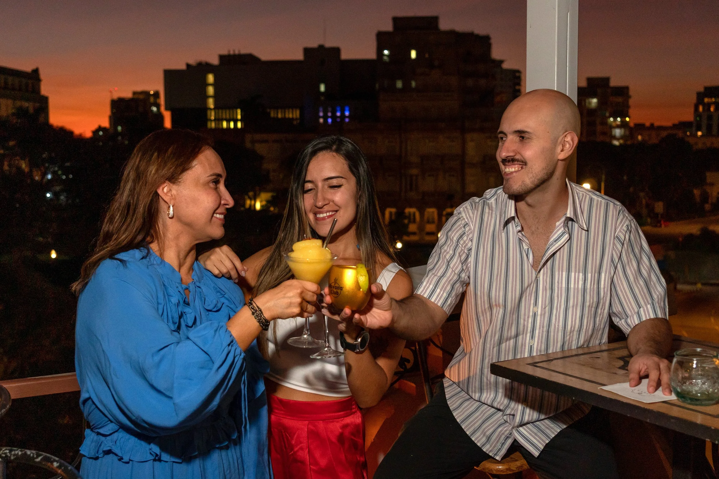 Three friends celebrating at an outdoor restaurant or bar during sunset, raising their drinks for a toast and smiling.
