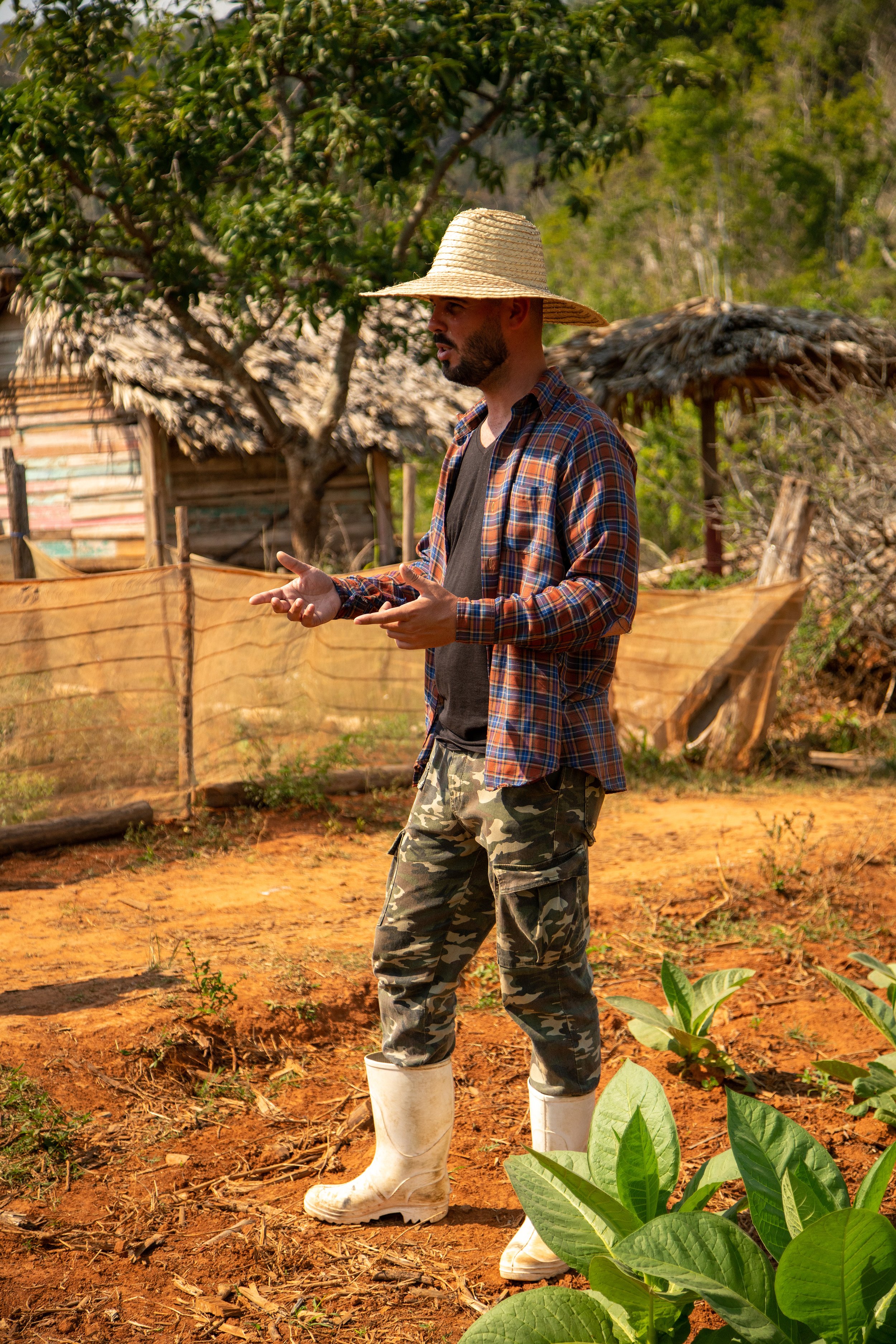 A man wearing a straw hat, checkered shirt, and camouflage pants with white rubber boots stands outdoors in a rural garden or farm, gesturing with his hands in Viñales, Cuba.