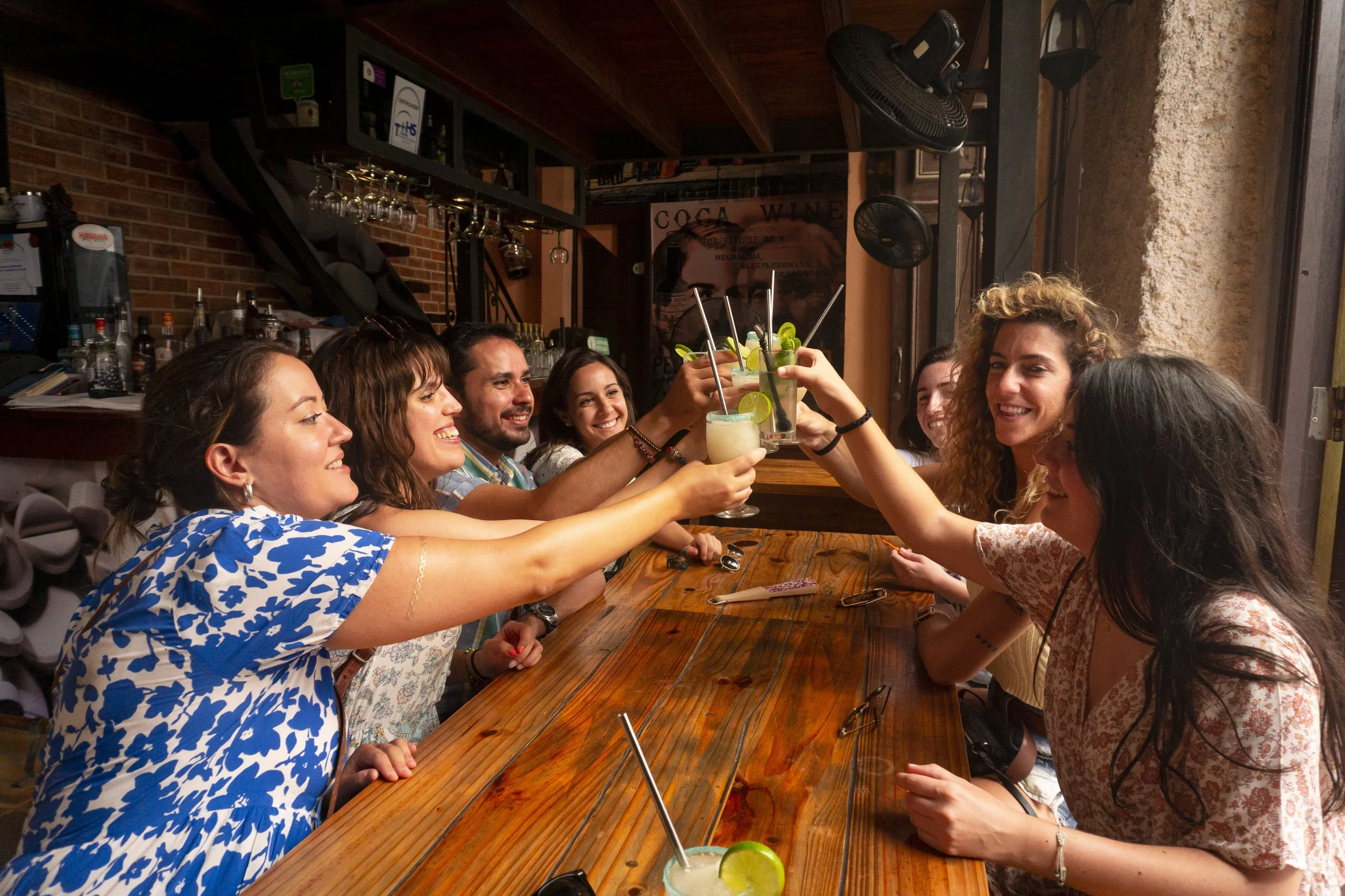 Group of seven friends sitting at a wooden bar, raising cocktails for a toast, smiling, in a lively, warmly lit bar or restaurant.