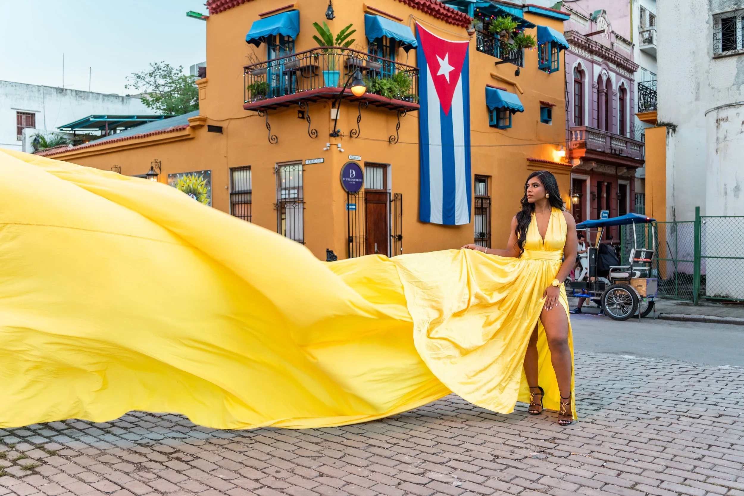 Woman in a long, flowing yellow dress standing on a cobblestone street in front of a colorful building with a large Cuban flag hanging outside.