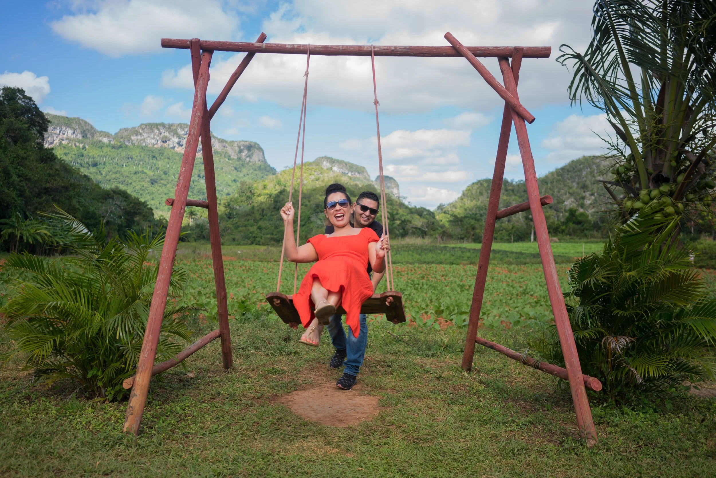 A joyful couple enjoying a swing ride outdoors in a lush green field with mountains in the background, during daytime with a partly cloudy sky.