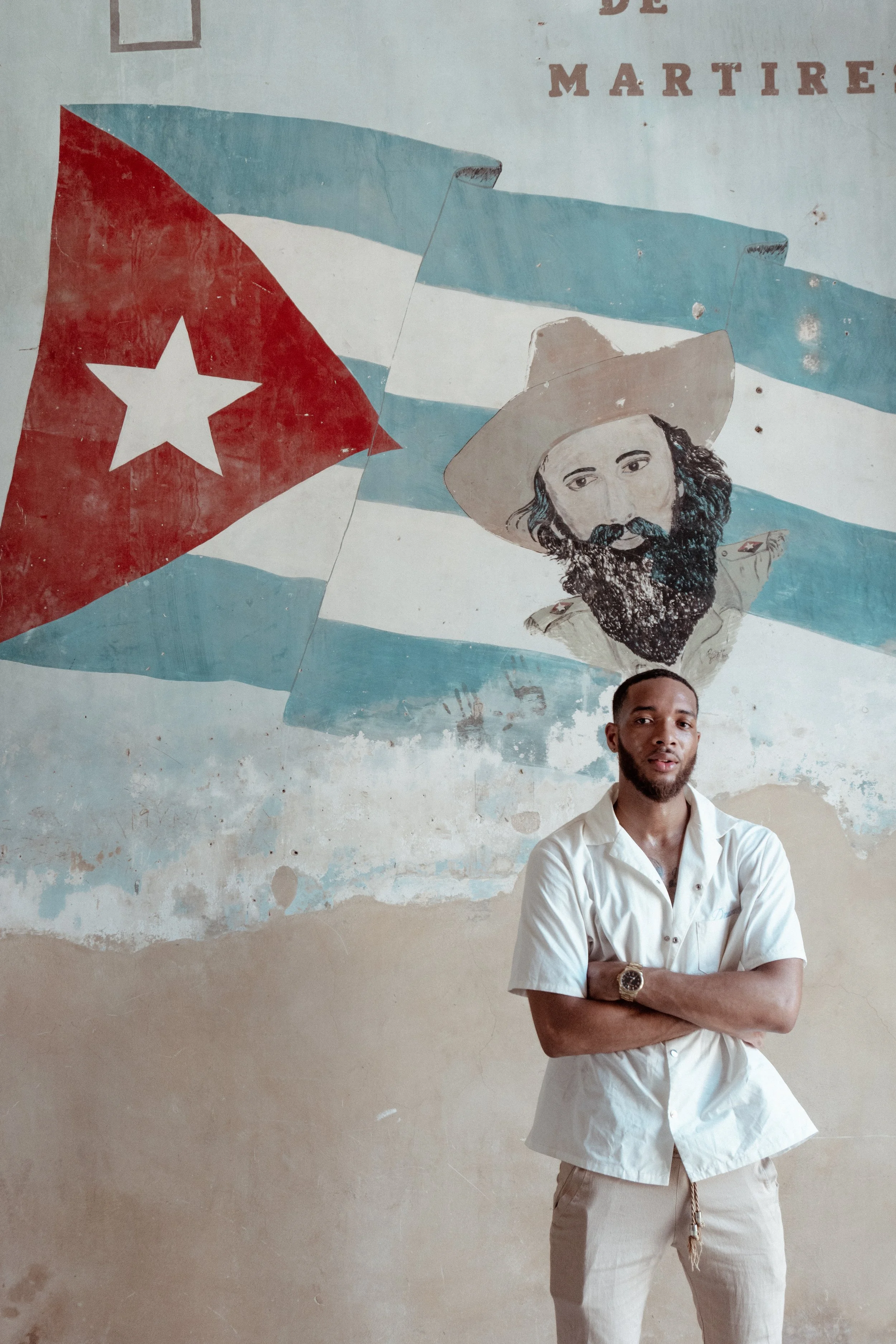 A man with crossed arms standing in front of a mural of the Cuban flag and a portrait of Fidel Castro on a weathered wall.