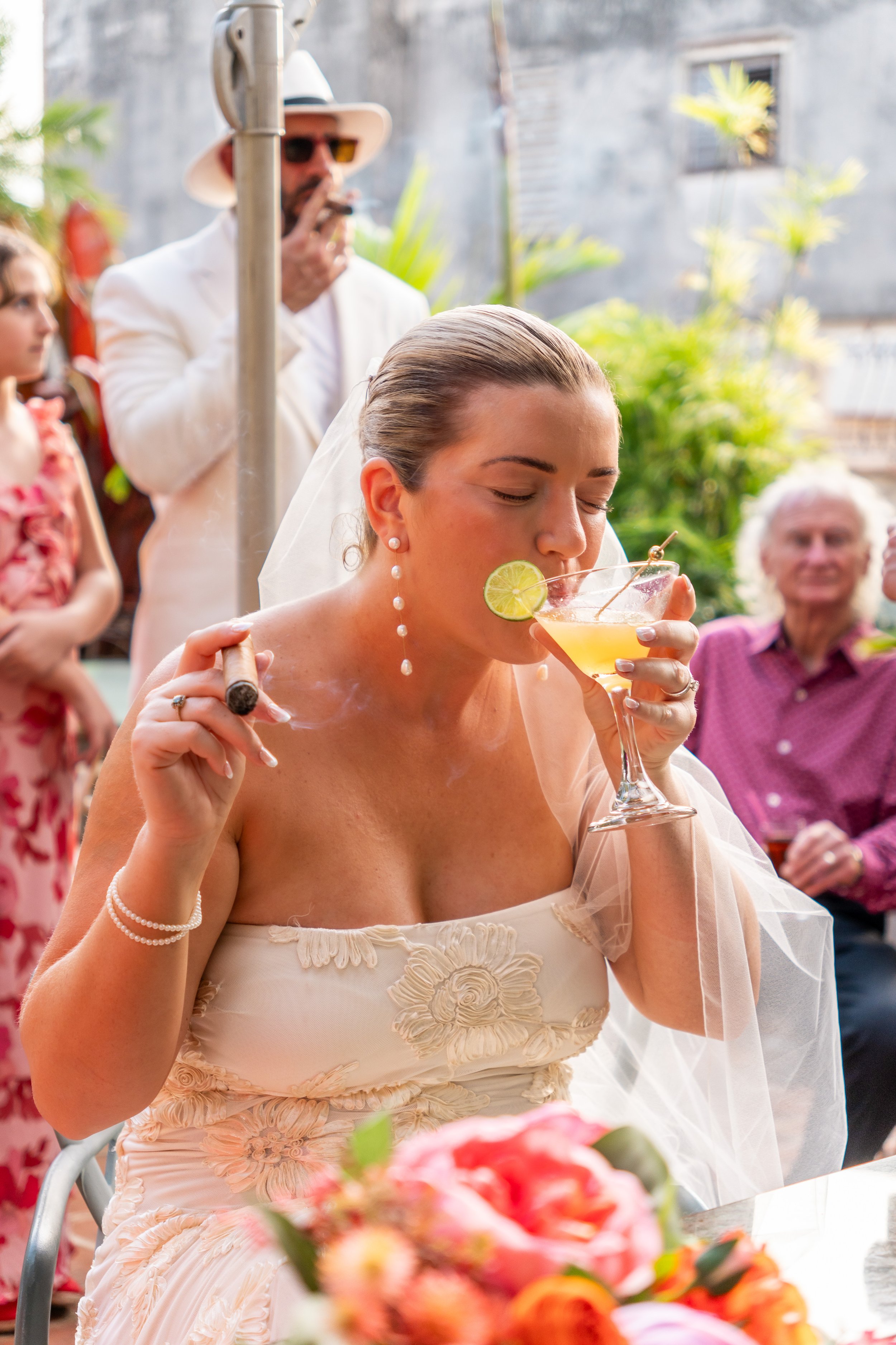 A bride with earrings and a veil is sitting outdoors, drinking a cocktail garnished with a lime slice and a cherry, while holding a lit cigar. People are gathered in the background at a social event with vibrant, tropical plants visible.