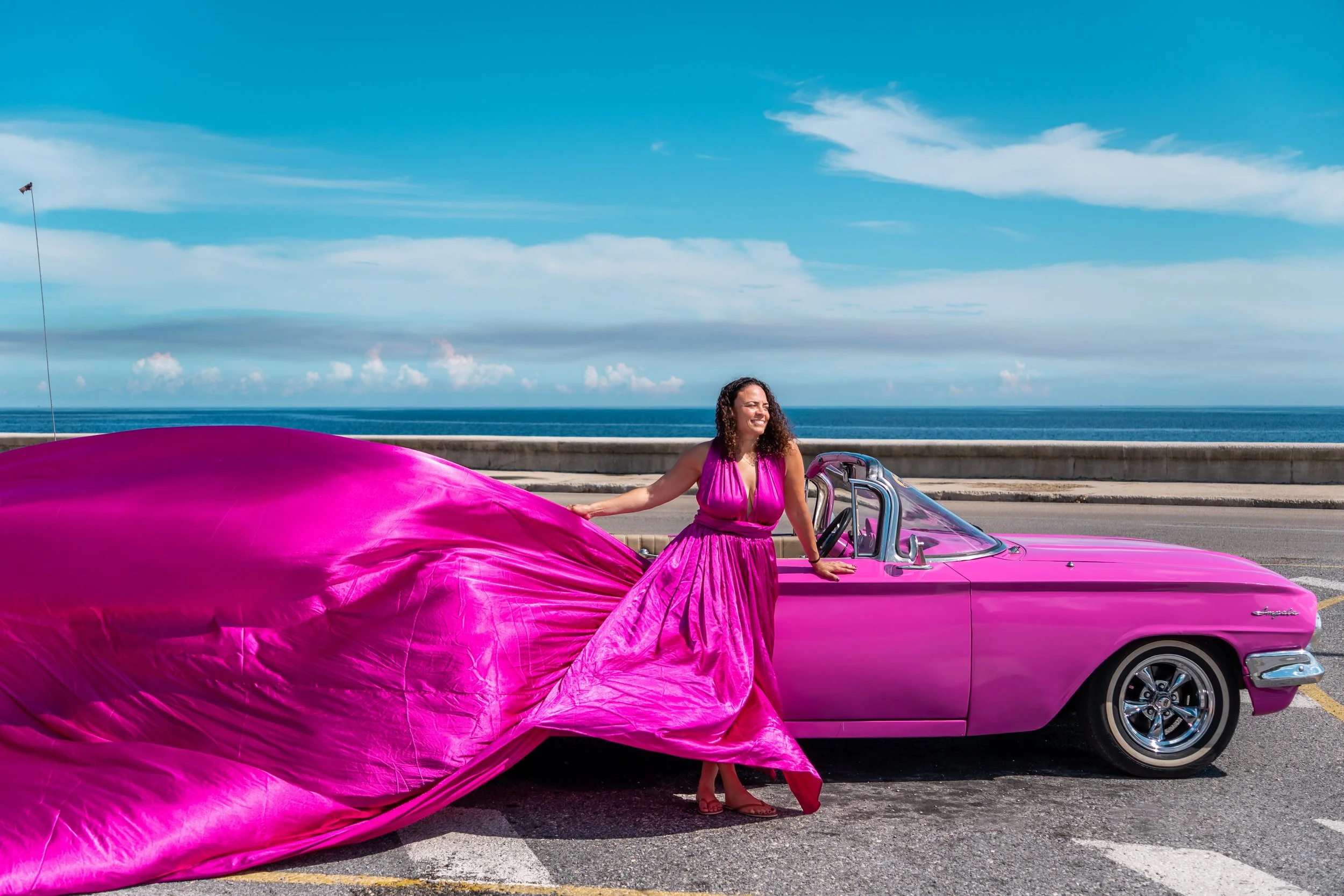 Woman in a pink dress standing beside a vintage pink convertible car on a coastal road with ocean and sky in the background.