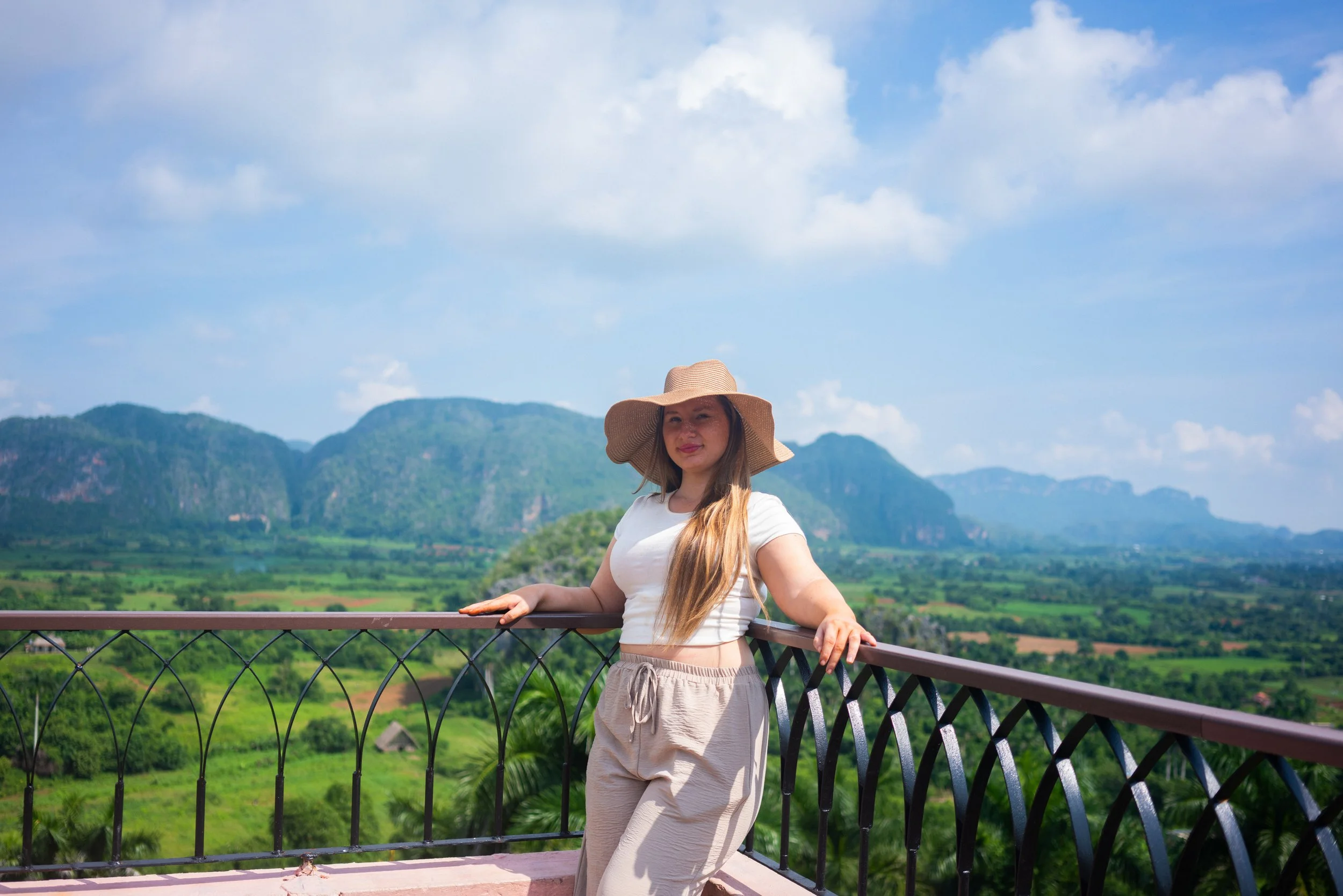 A woman in a white crop top, beige pants, and a wide-brimmed hat stands on a balcony with a scenic mountain landscape in the background, under a partly cloudy sky.