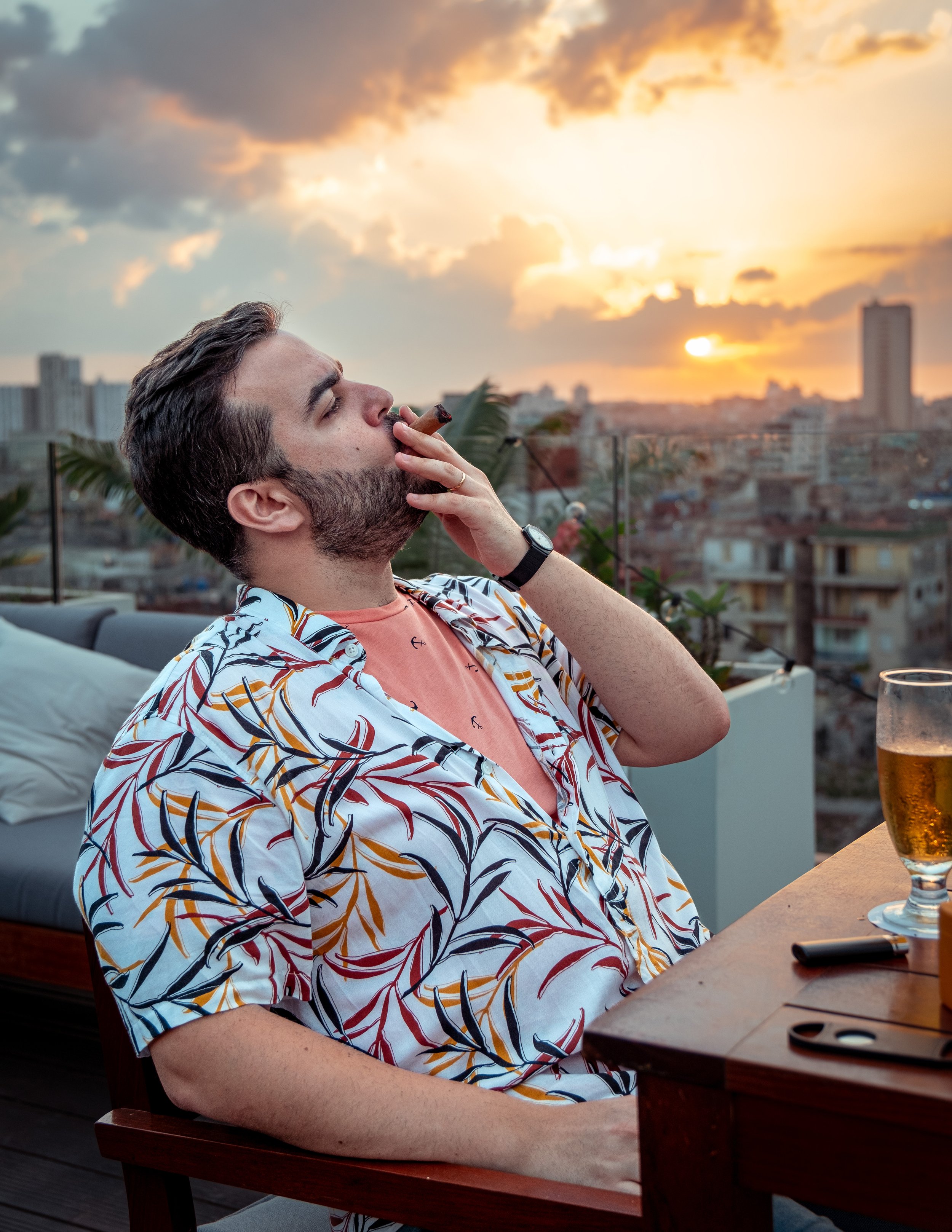 A man with a beard, wearing a floral shirt and pink t-shirt, sitting outdoors at sunset, smoking a cigar and holding a glass of beer.