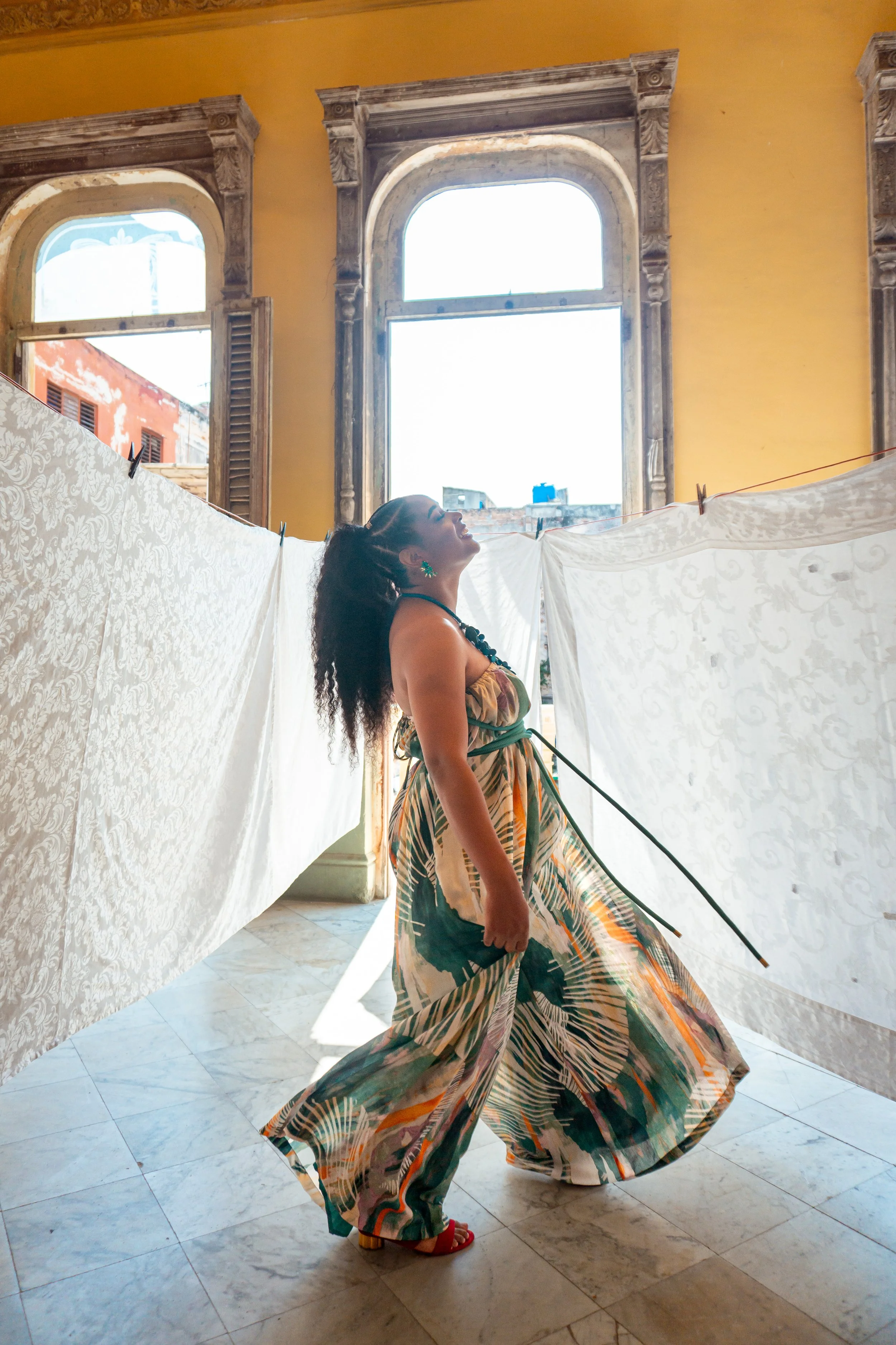 A woman in a colorful, flowing dress stands inside a room with large windows and ornate window frames. She is posing with her head tilted back, surrounded by white patterned fabric or sheets hanging on a line. The sunlight streams through the window,