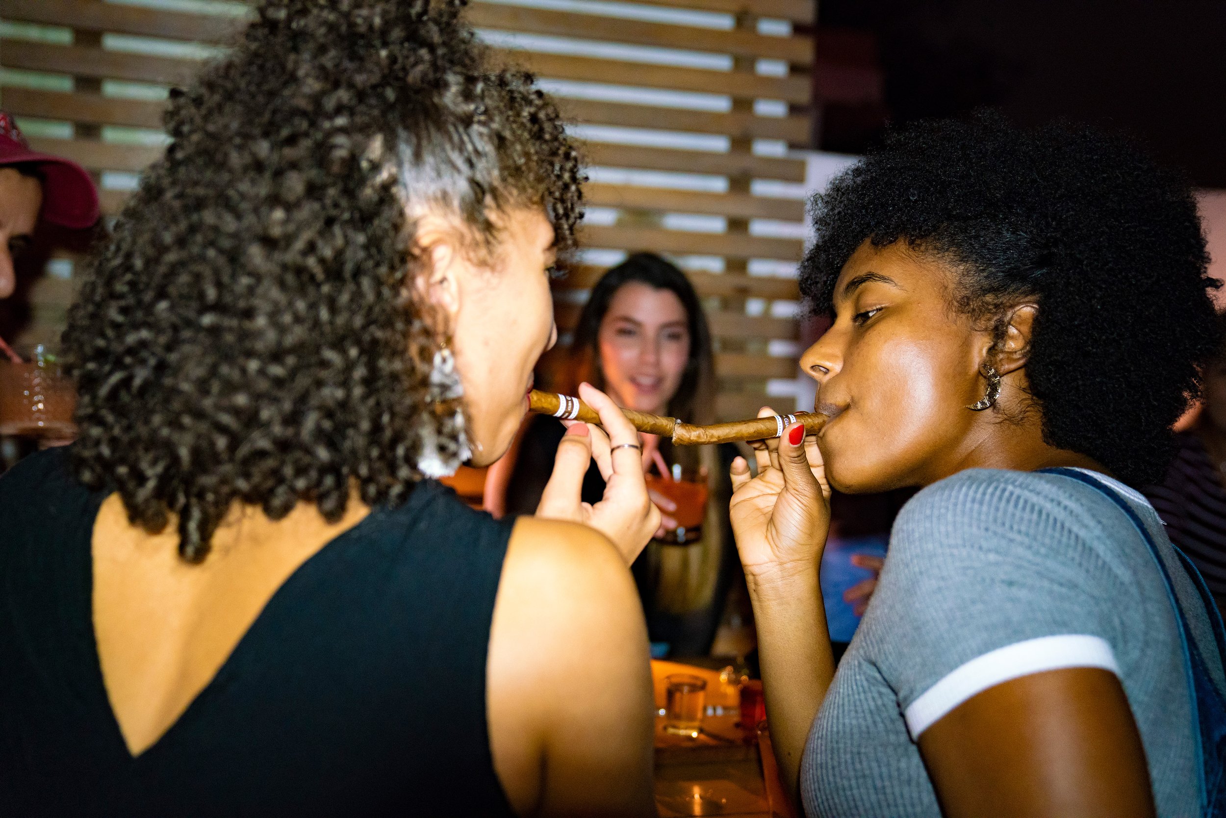 Two women sharing a cigar, with one woman handing the cigar to the other, while a woman in the background looks on smiling in a social setting.