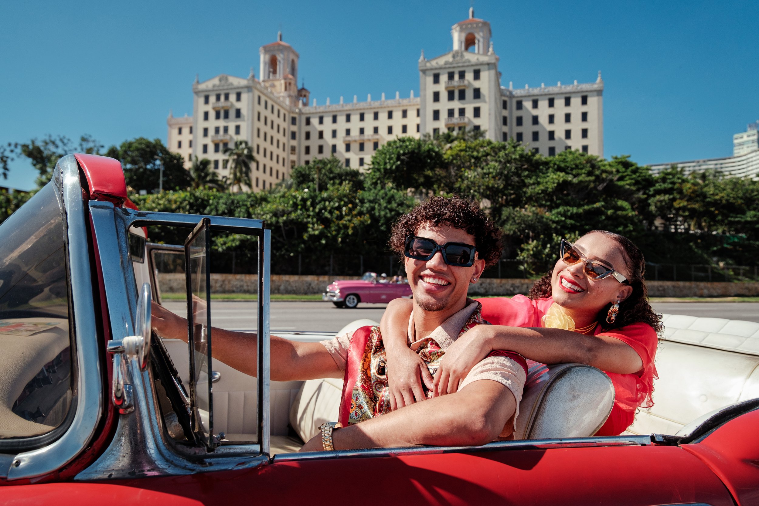 A smiling couple enjoying a convertible ride in front of a large historic hotel with a lush green hill and clear blue sky in the background.