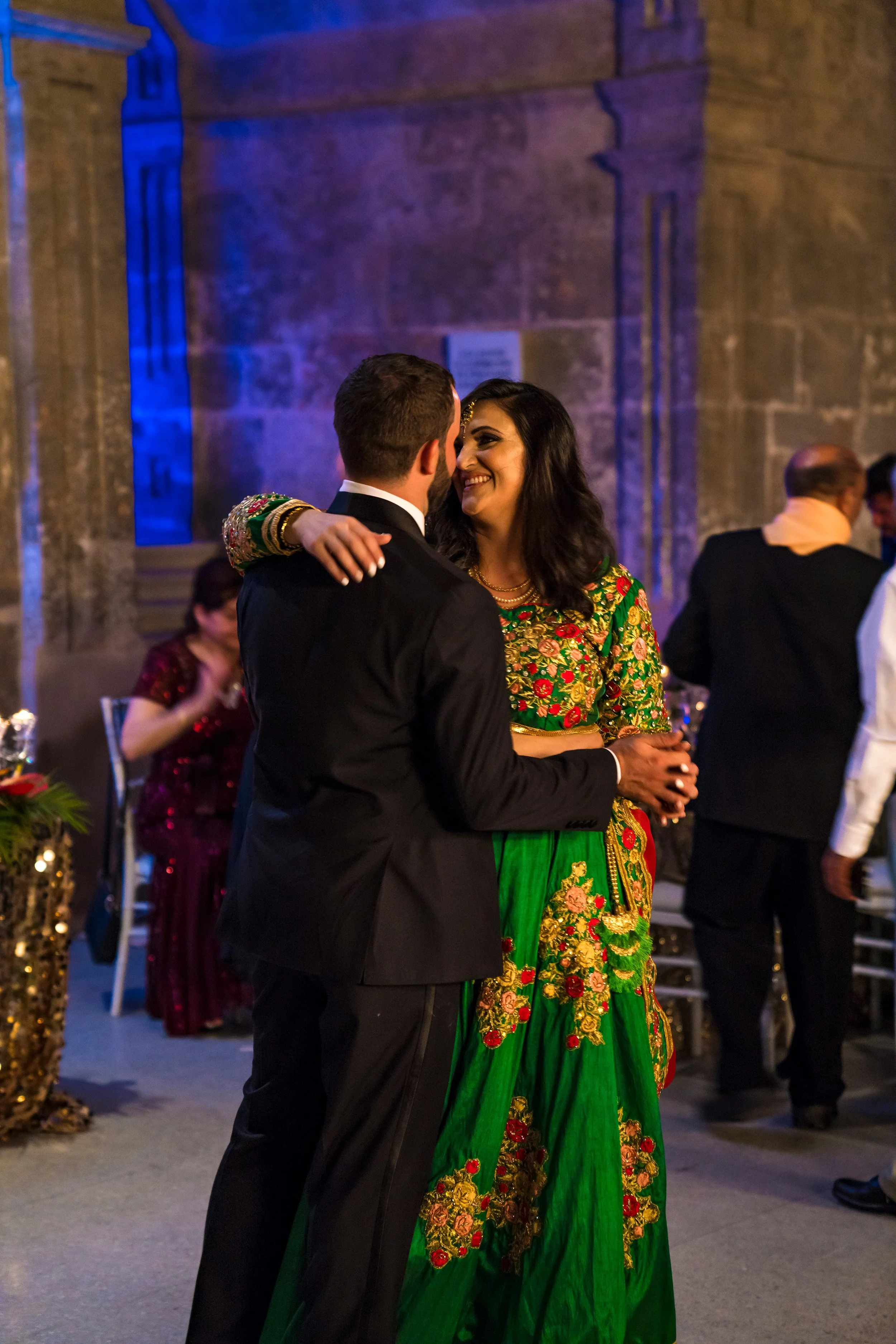 A couple dancing at a formal event, dressed in traditional South Asian attire, with the woman wearing a vibrant green and gold embroidered outfit and the man in a black suit, inside a decorated venue with other guests in the background.
