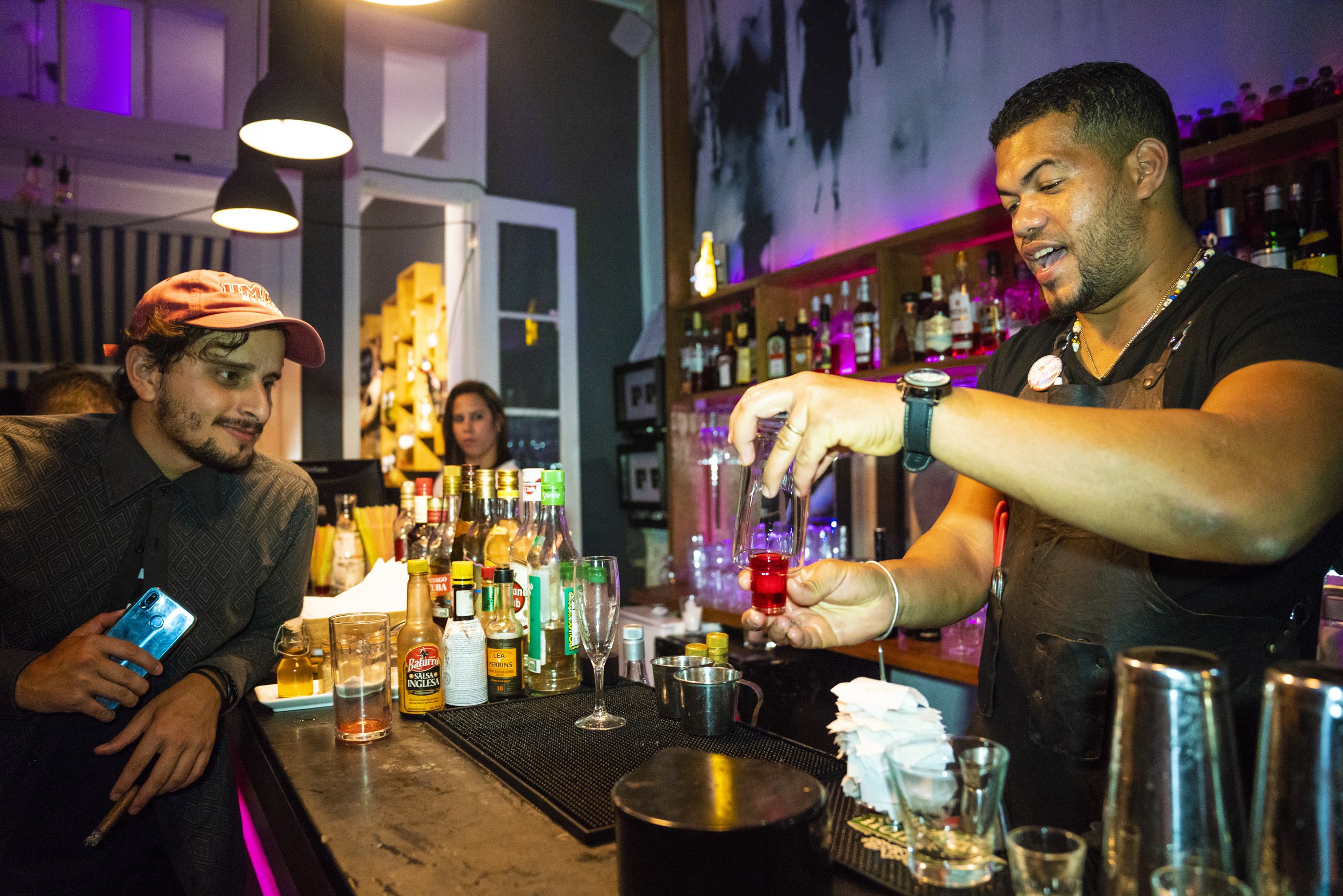 A bartender preparing a red drink at a bar counter while a customer, holding a phone, watches. The bartender is pouring a red liquid into a glass, with various liquor bottles on shelves behind him. Purple and blue ambient lighting highlights the scen