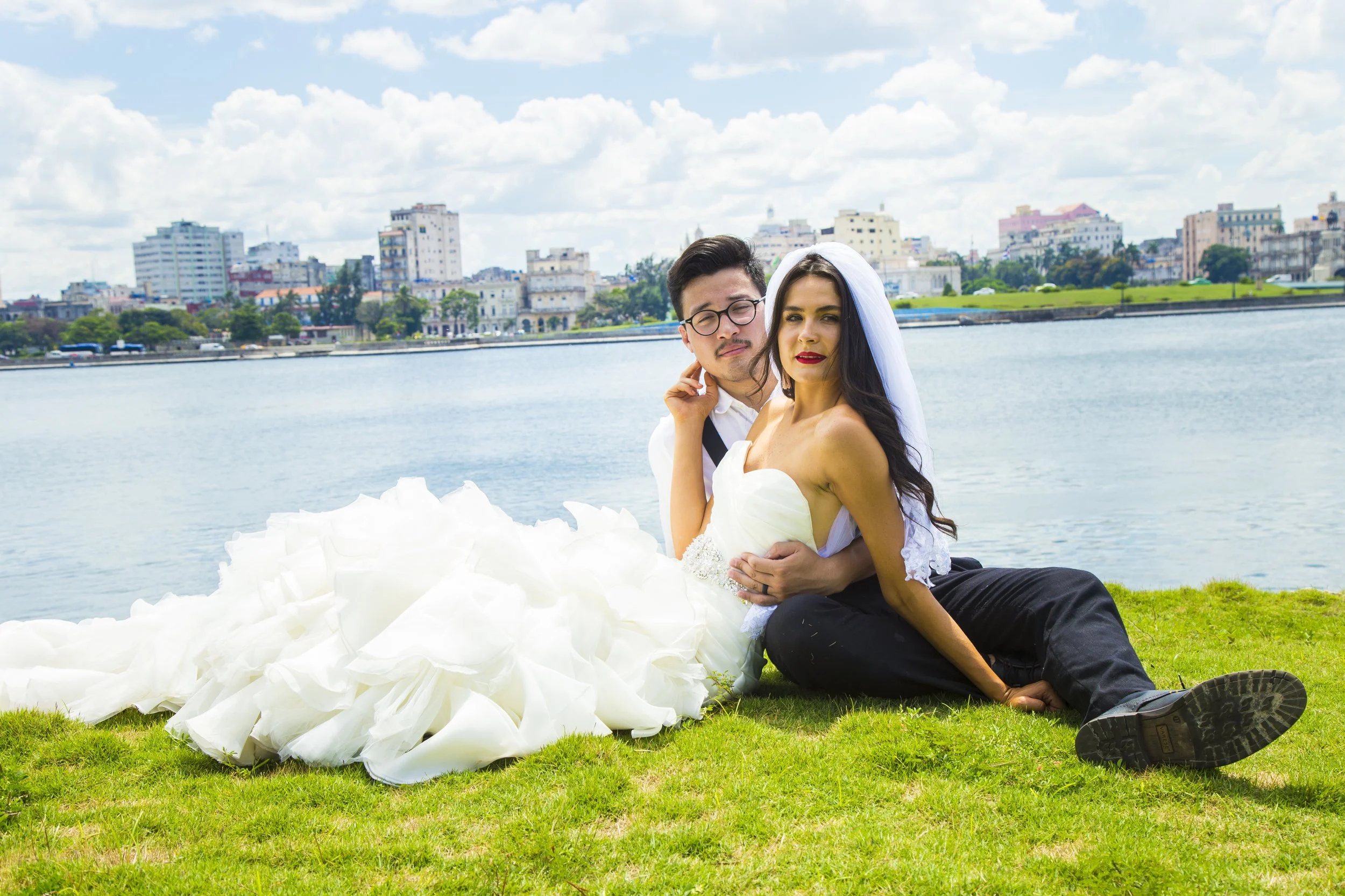 A bride and groom sitting on grass by a body of water with a city skyline in the background. The bride is wearing a white wedding dress with a veil, and the groom is in black pants and a white shirt. They are close together, with the groom holding th