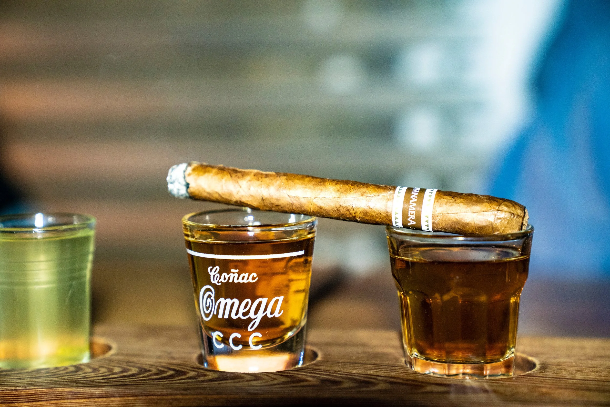 Three shot glasses with brown liquor on a wooden surface, with a lit cigar resting on the rims of two glasses. The background is blurred.