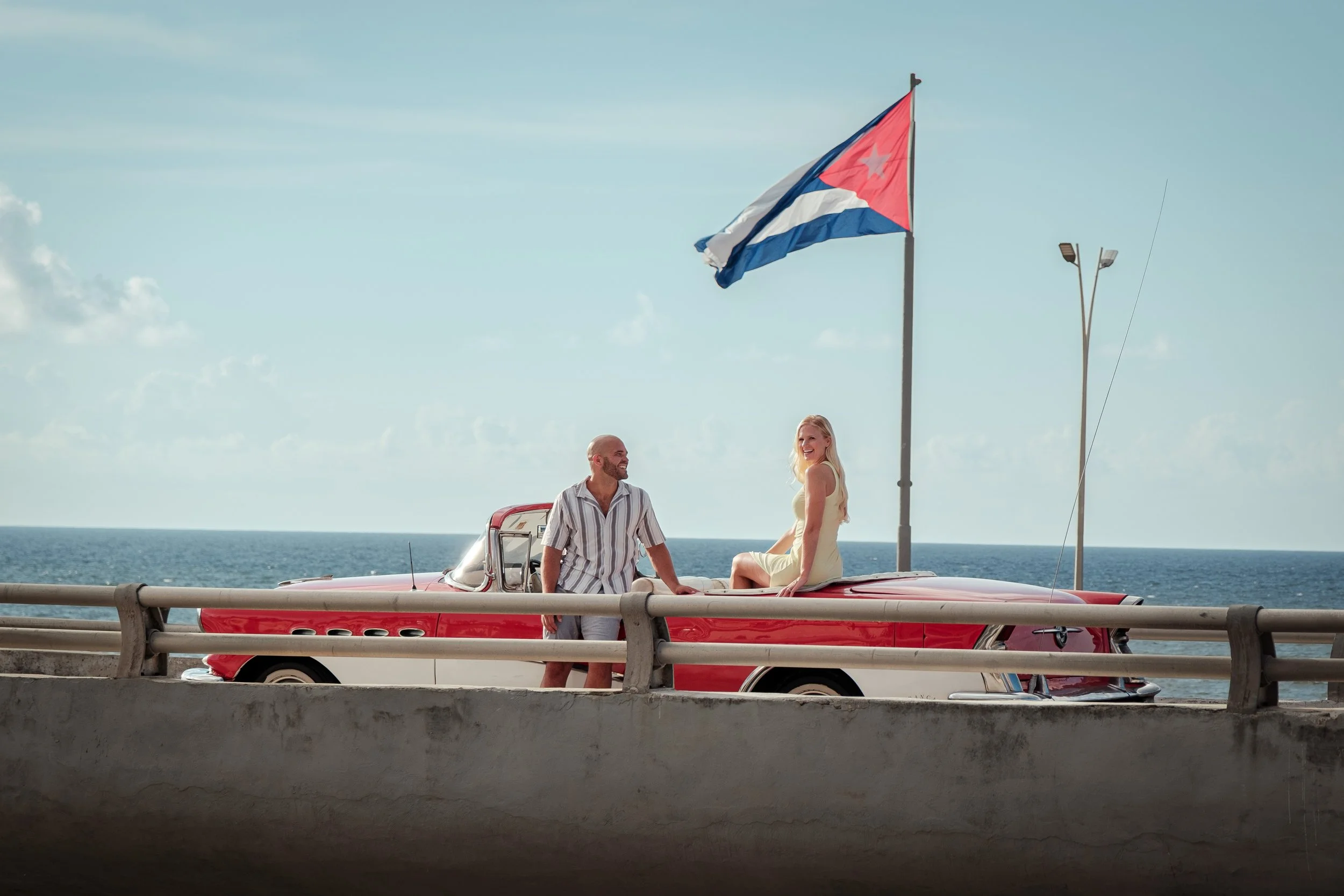 A man and woman enjoying a day by the ocean, sitting on a vintage red and white car, with a Cuban flag flying in the background.