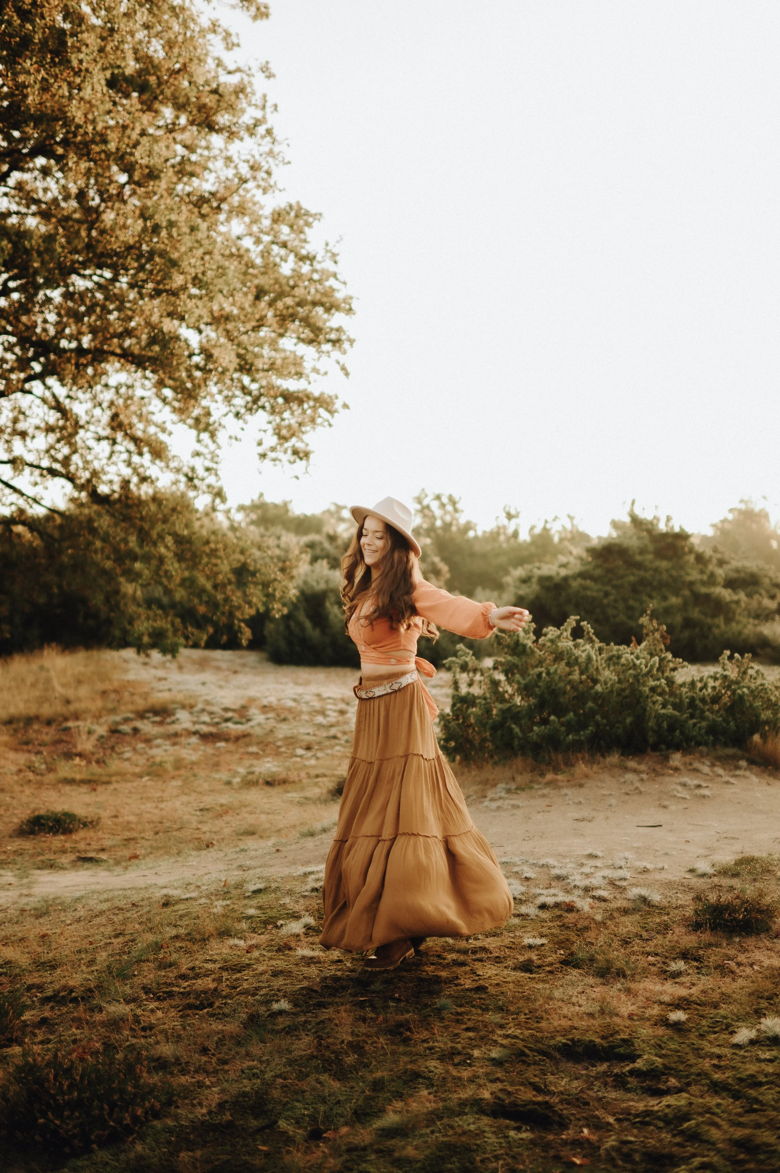 Vrouw die danst in een veld, gekleed in een bruinrokken en een lichte jurk, met een hoed op haar hoofd, met bomen en struiken op de achtergrond tijdens zonsondergang.