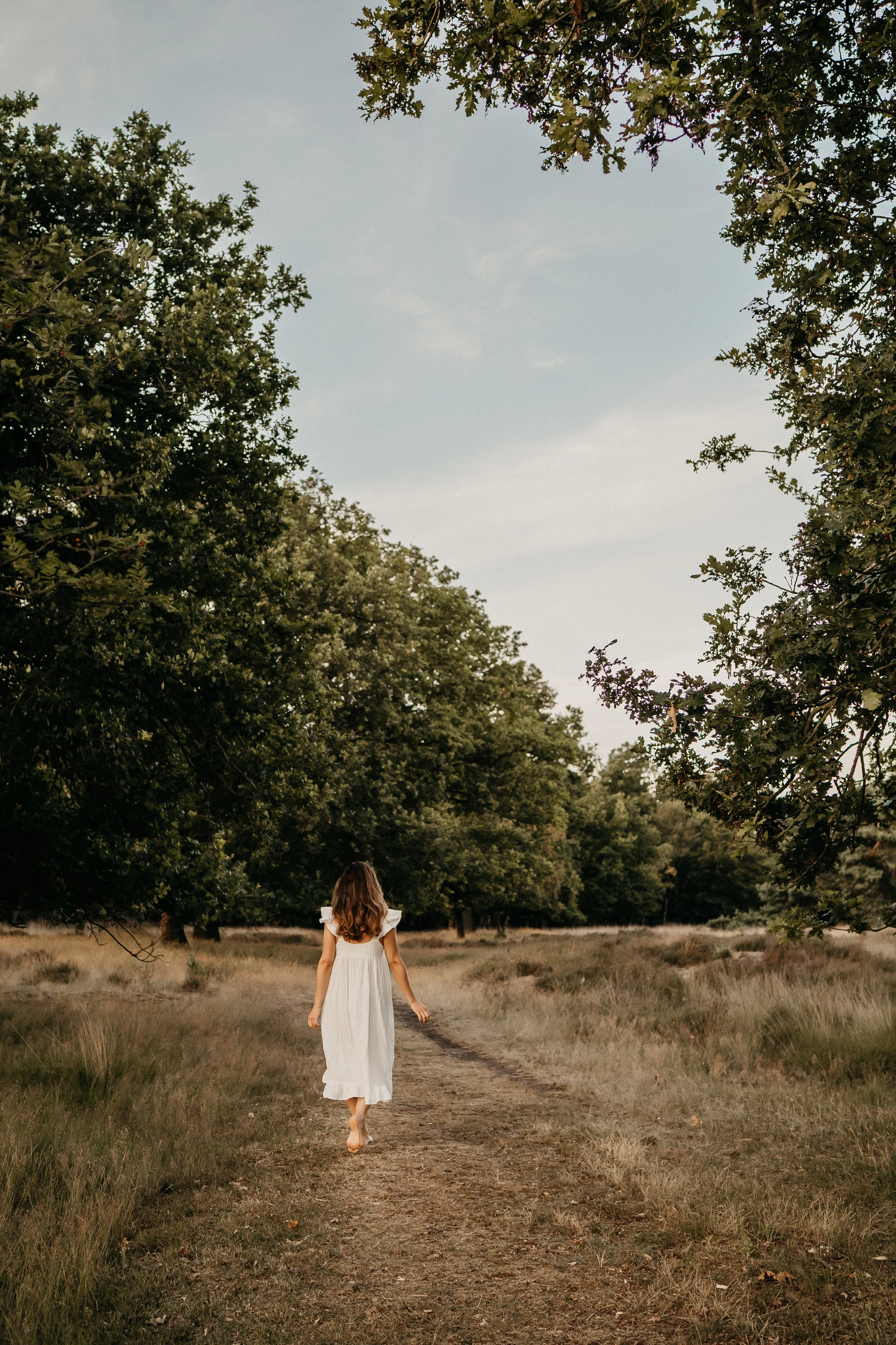 Een meisje in een witte jurk loopt op een smal bospaadje, omgeven door gras en bomen, onder een blauwe lucht.