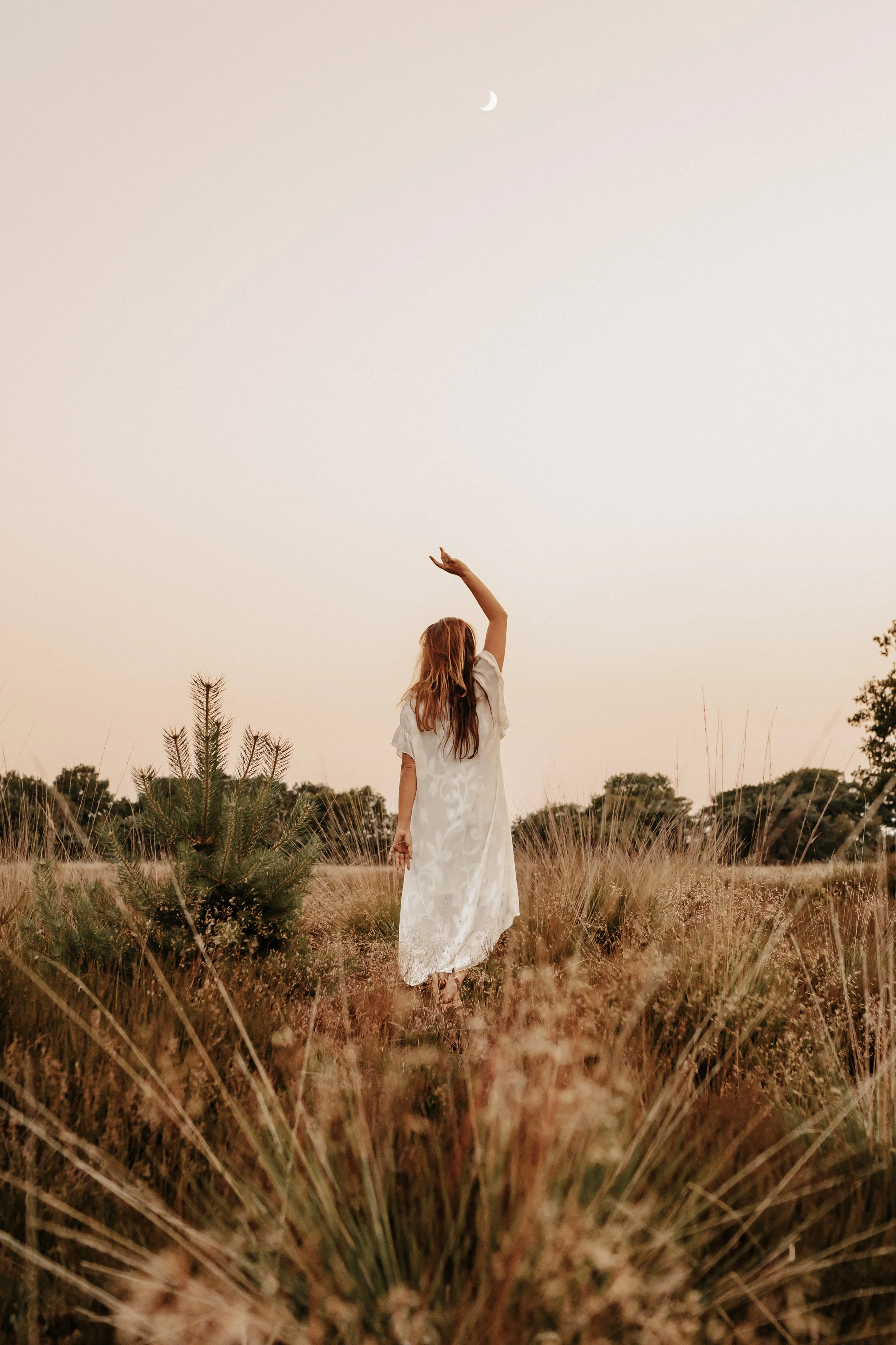 Vrouw in witte jurk in een koele veld, wekt de maan op en wijst ernaar.