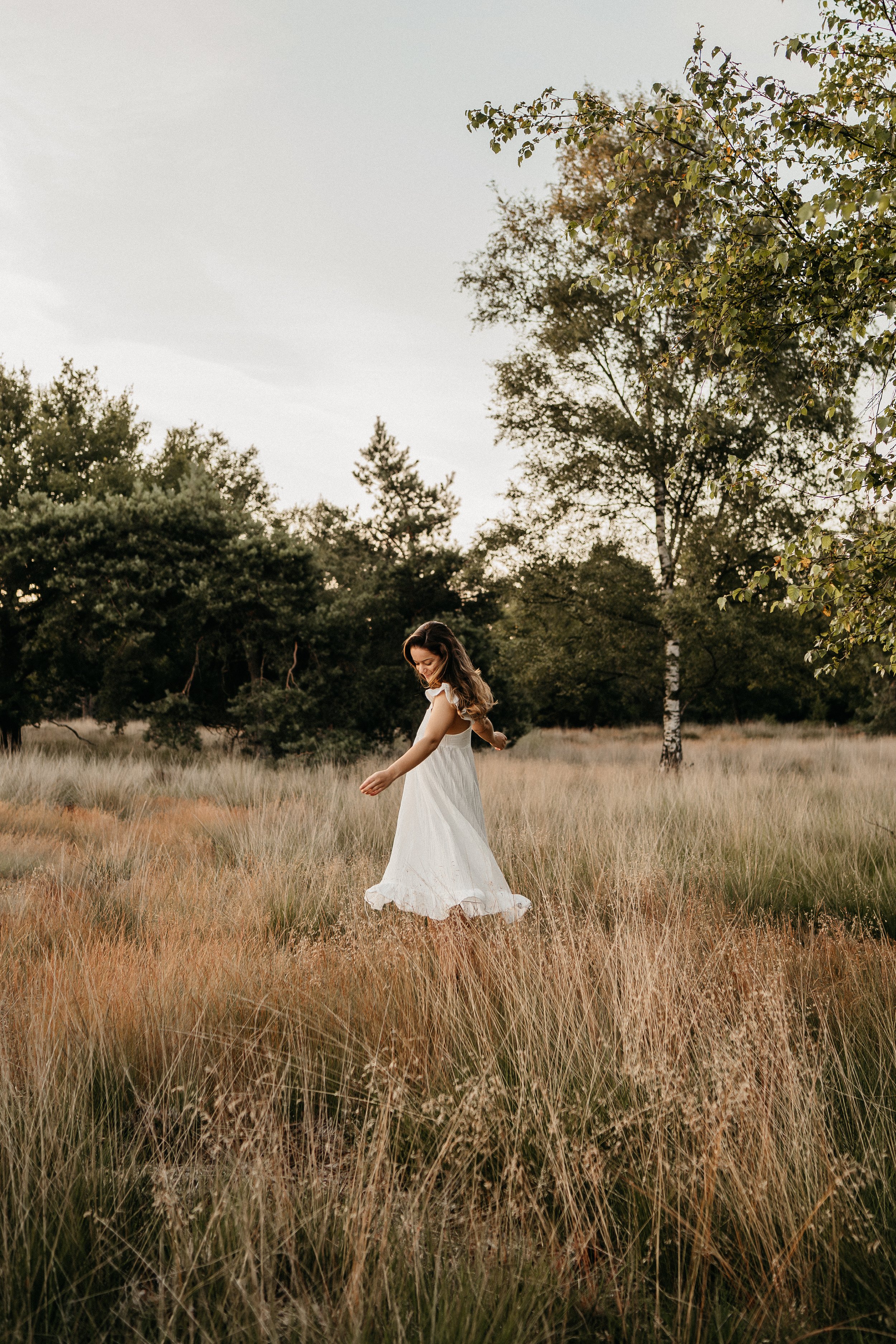 Een vrouw in een lange witte jurk die door een grasachtig veld in de natuur wandelt.